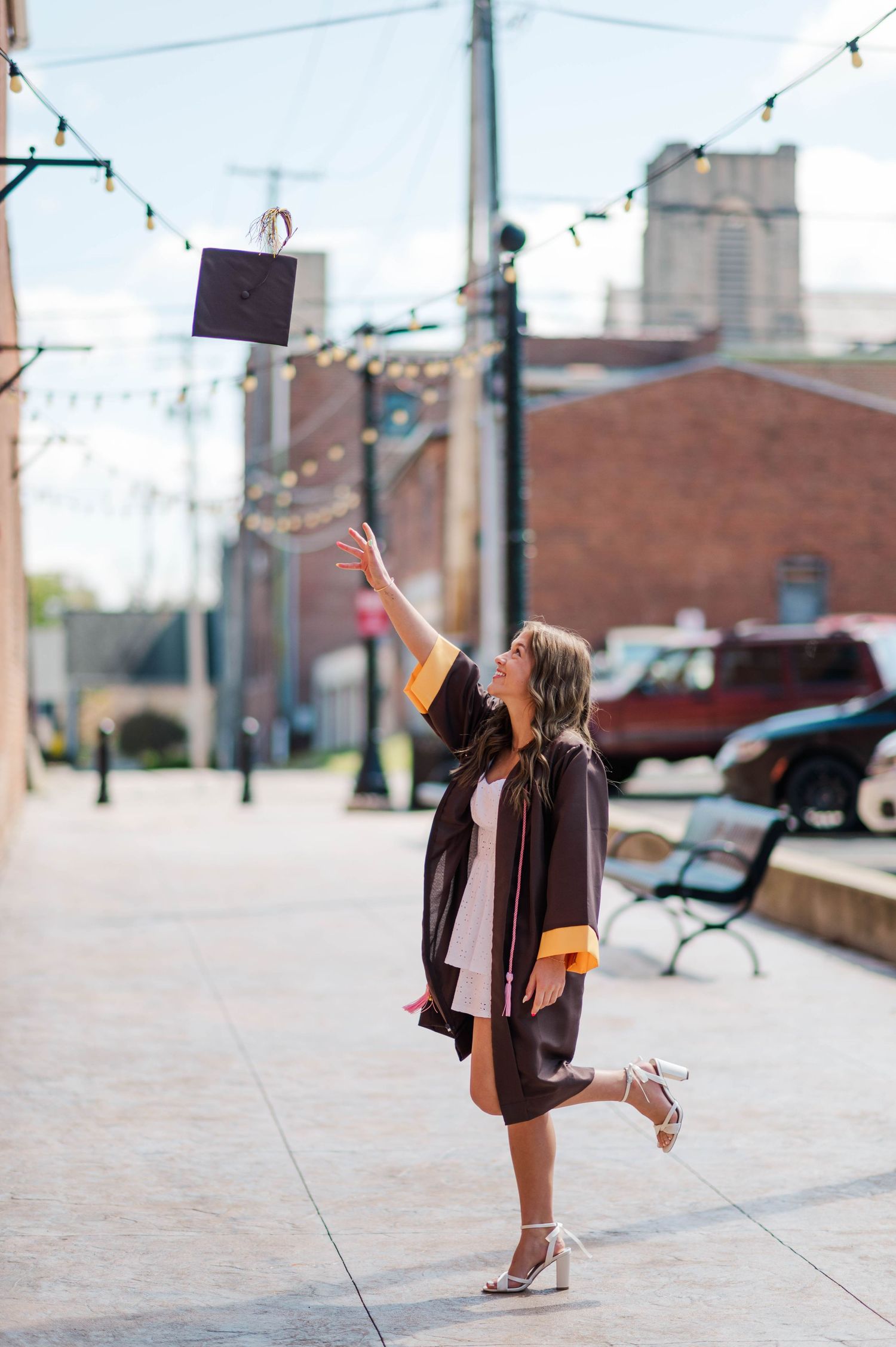 A smiling graduate in black and gold gown throws her cap skyward on a bright city sidewalk.