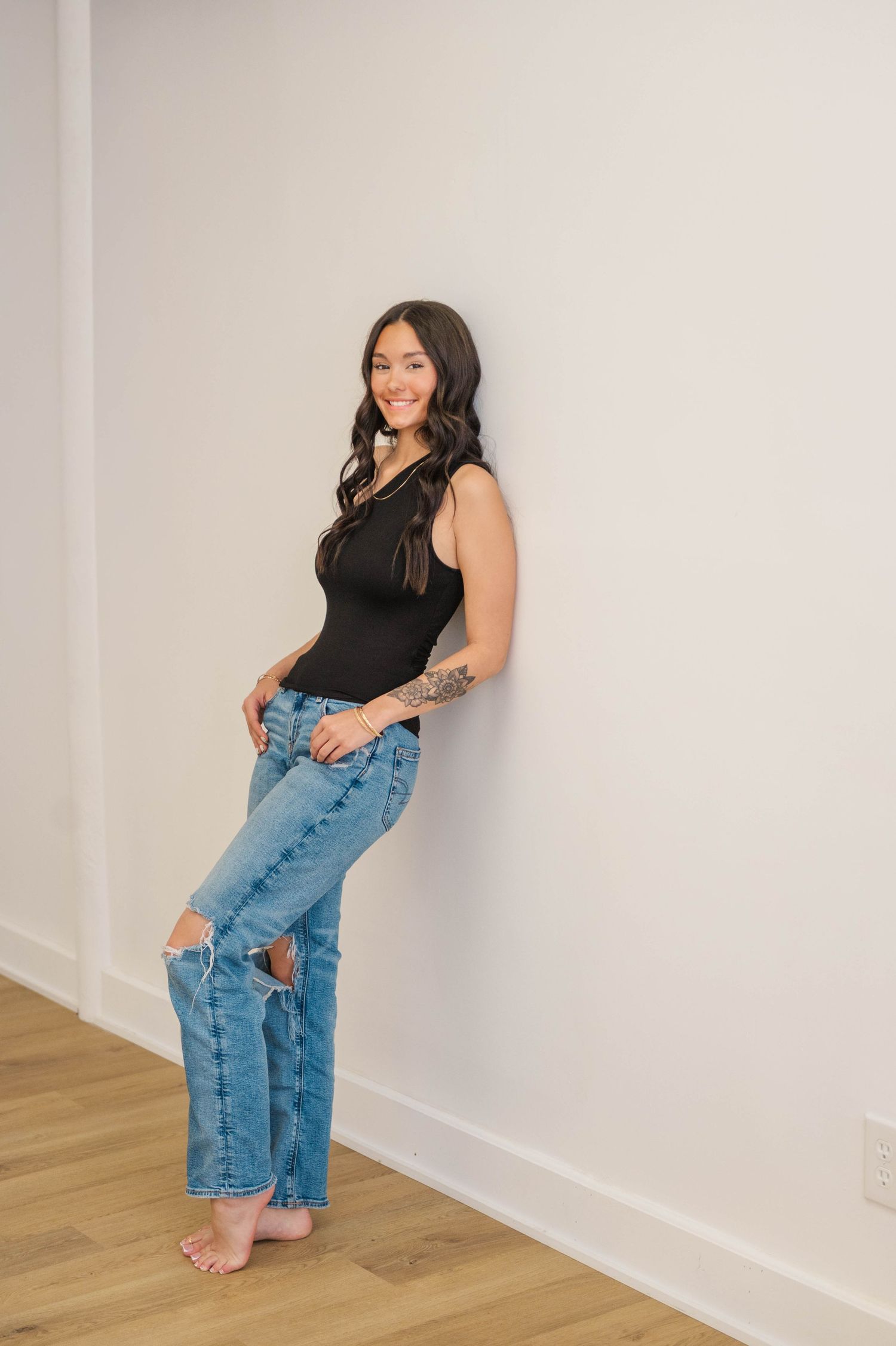 Smiling brunette woman with tattoos leans against white wall in black bodysuit and ripped jeans.