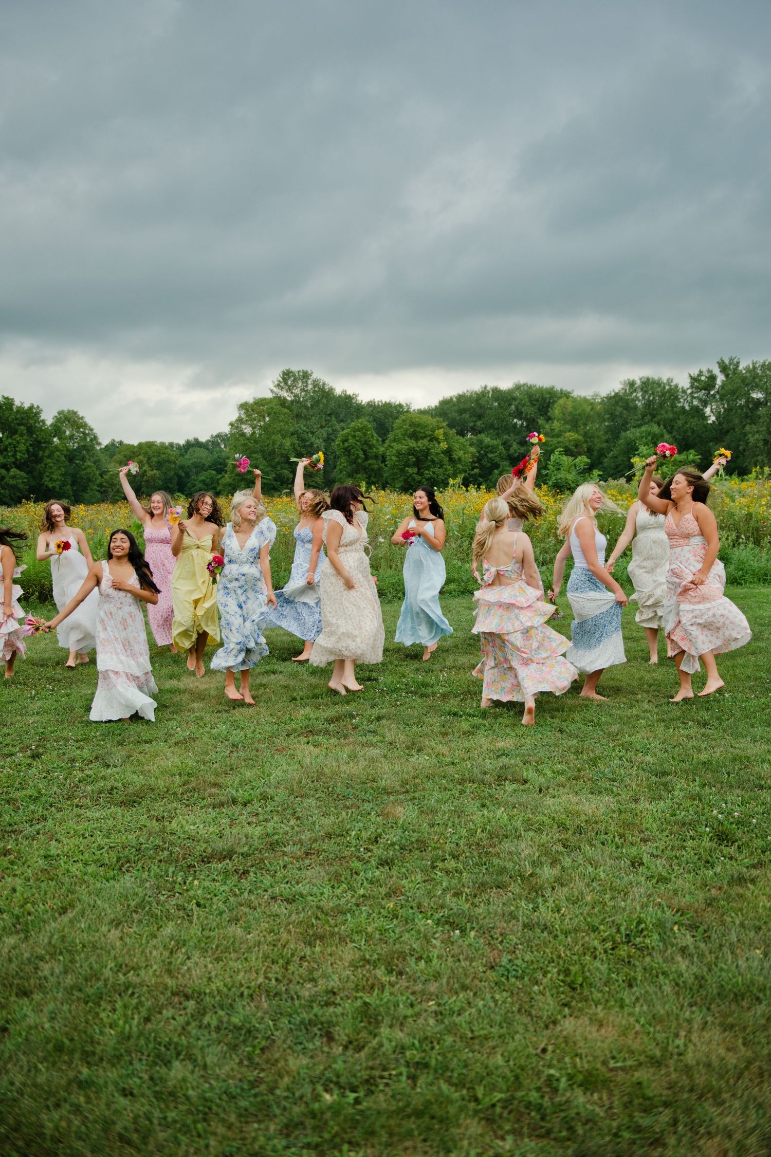Bridesmaids in colorful mismatched dresses dancing and twirling joyfully in a green field on an overcast day.