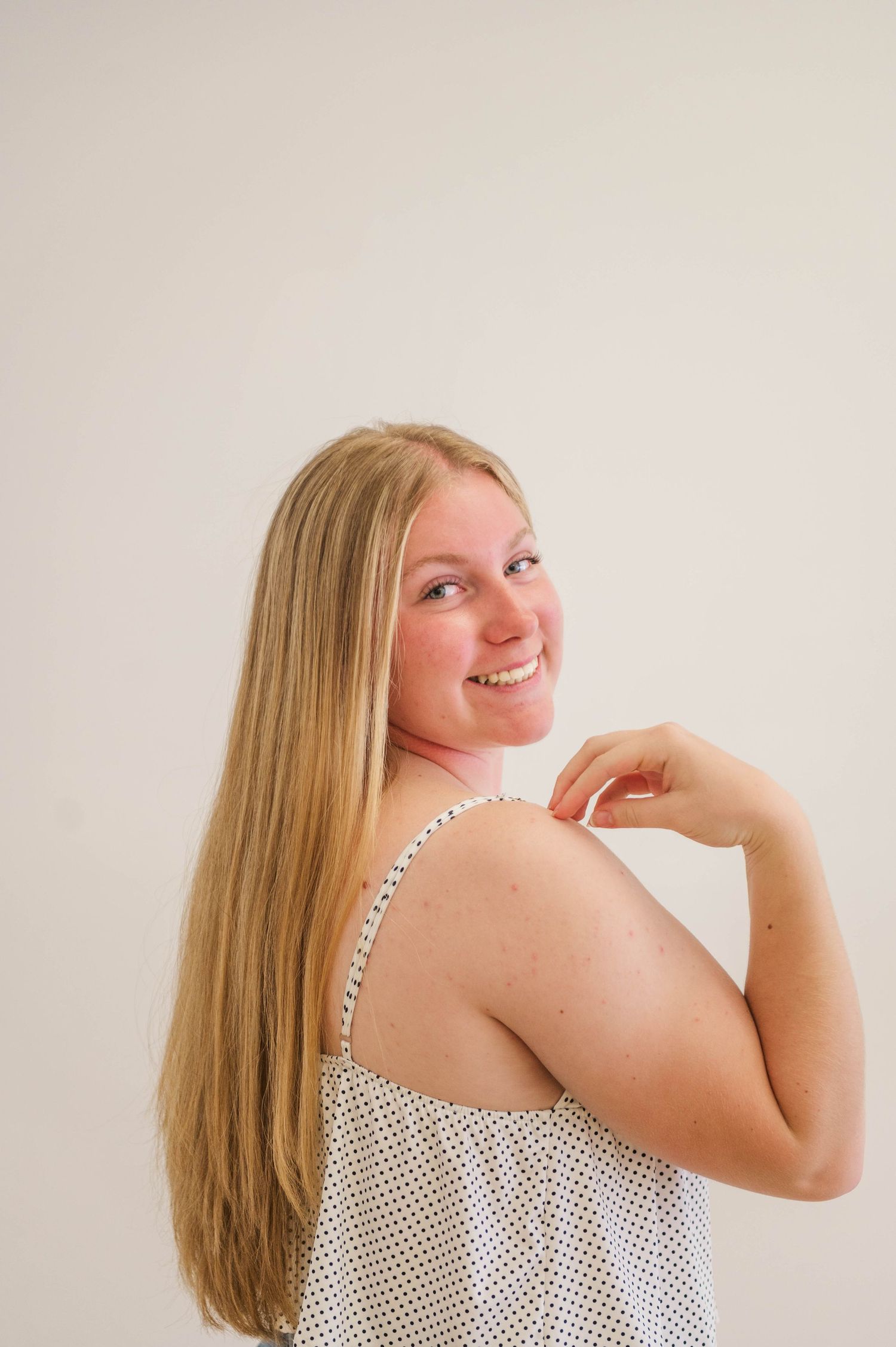 Smiling blonde woman in polka dot tank top looking over shoulder against white background.