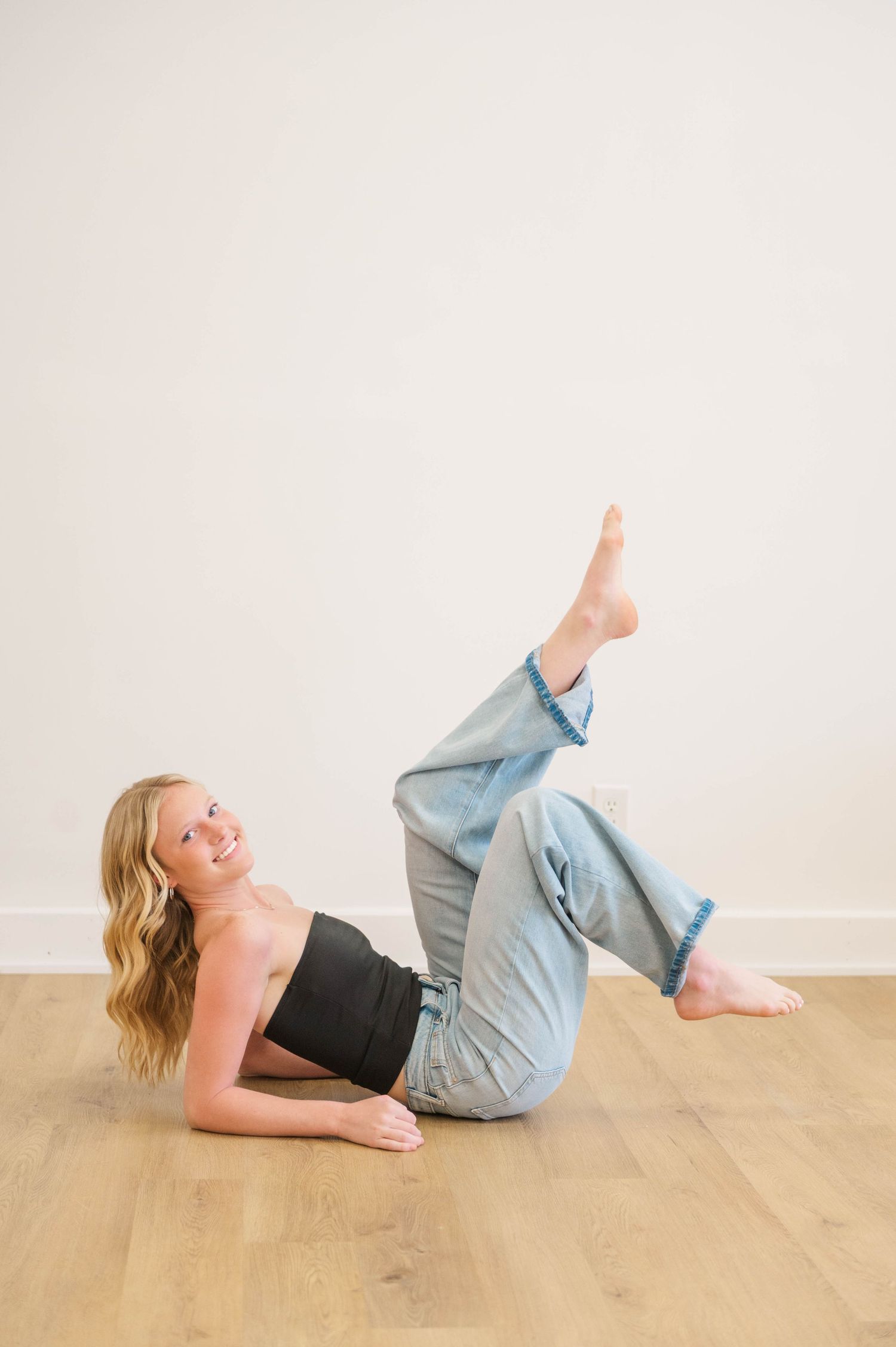 Smiling blonde woman reclines on floor in black tube top and light blue jeans with legs raised playfully.