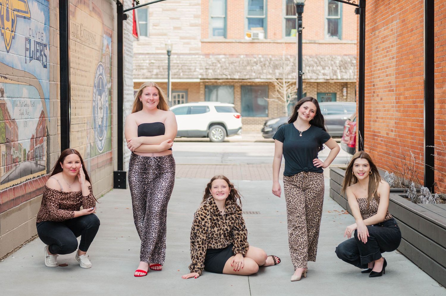 Six young women posing playfully on a downtown sidewalk, one seated, wearing coordinated leopard and black outfits.