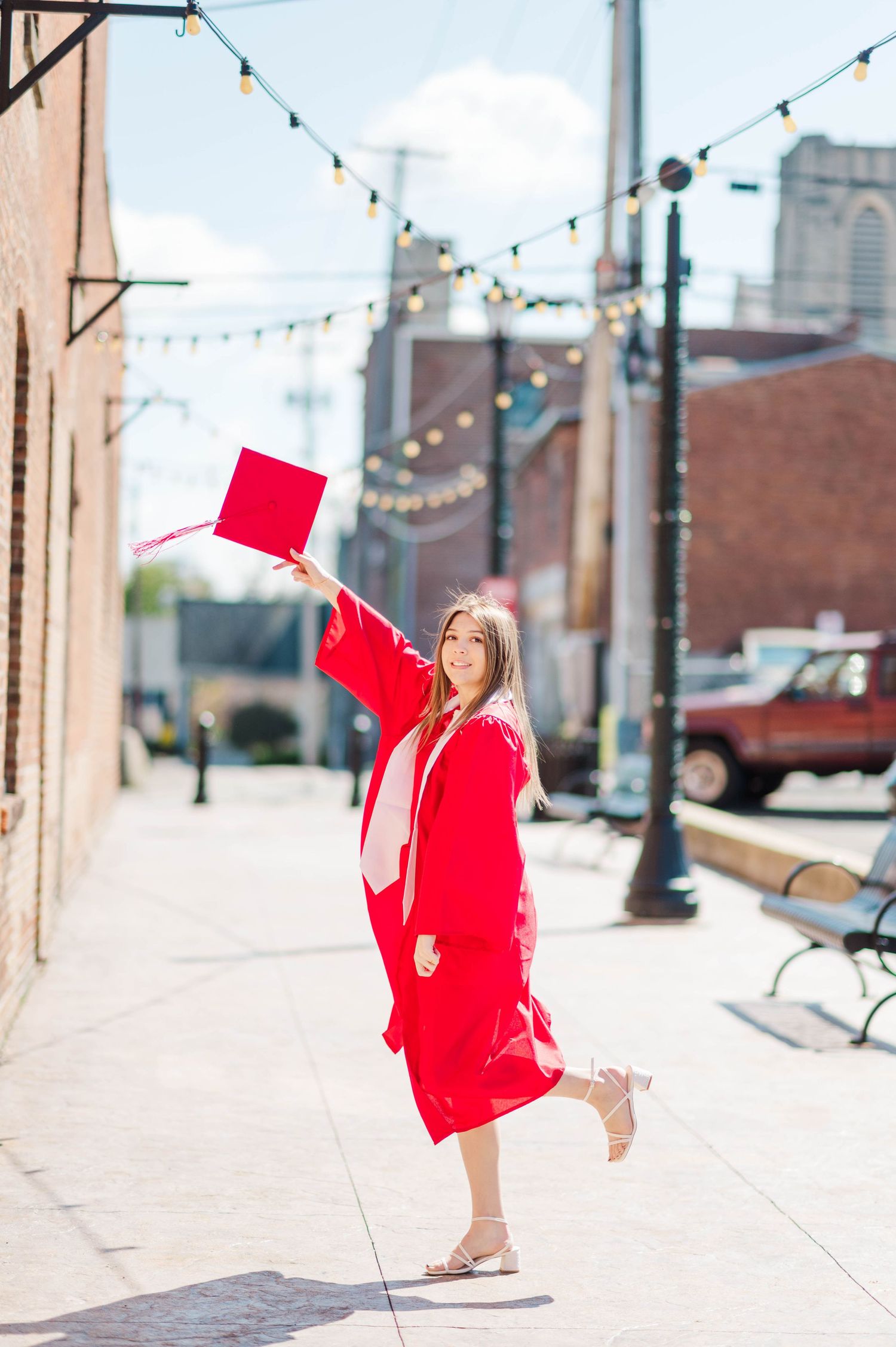 Smiling graduate in red cap and gown celebrating outdoors on a sunny city sidewalk.
