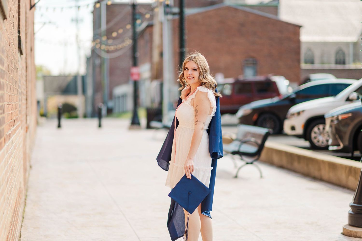 Smiling graduate in pink dress carries navy cap and gown along sunny brick downtown street.