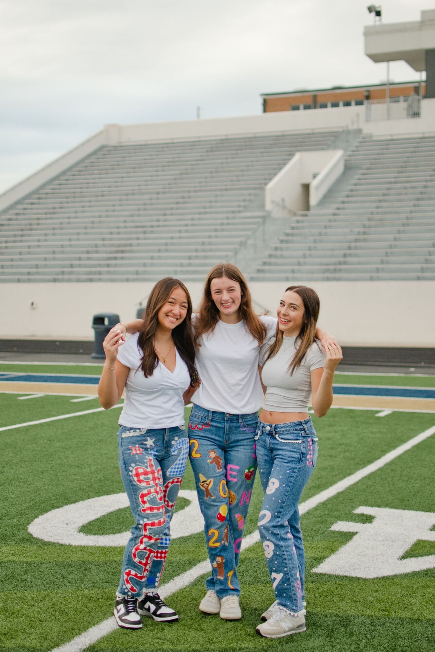 Three girls in white tops and colorful painted senior jeans smile together on a football field.