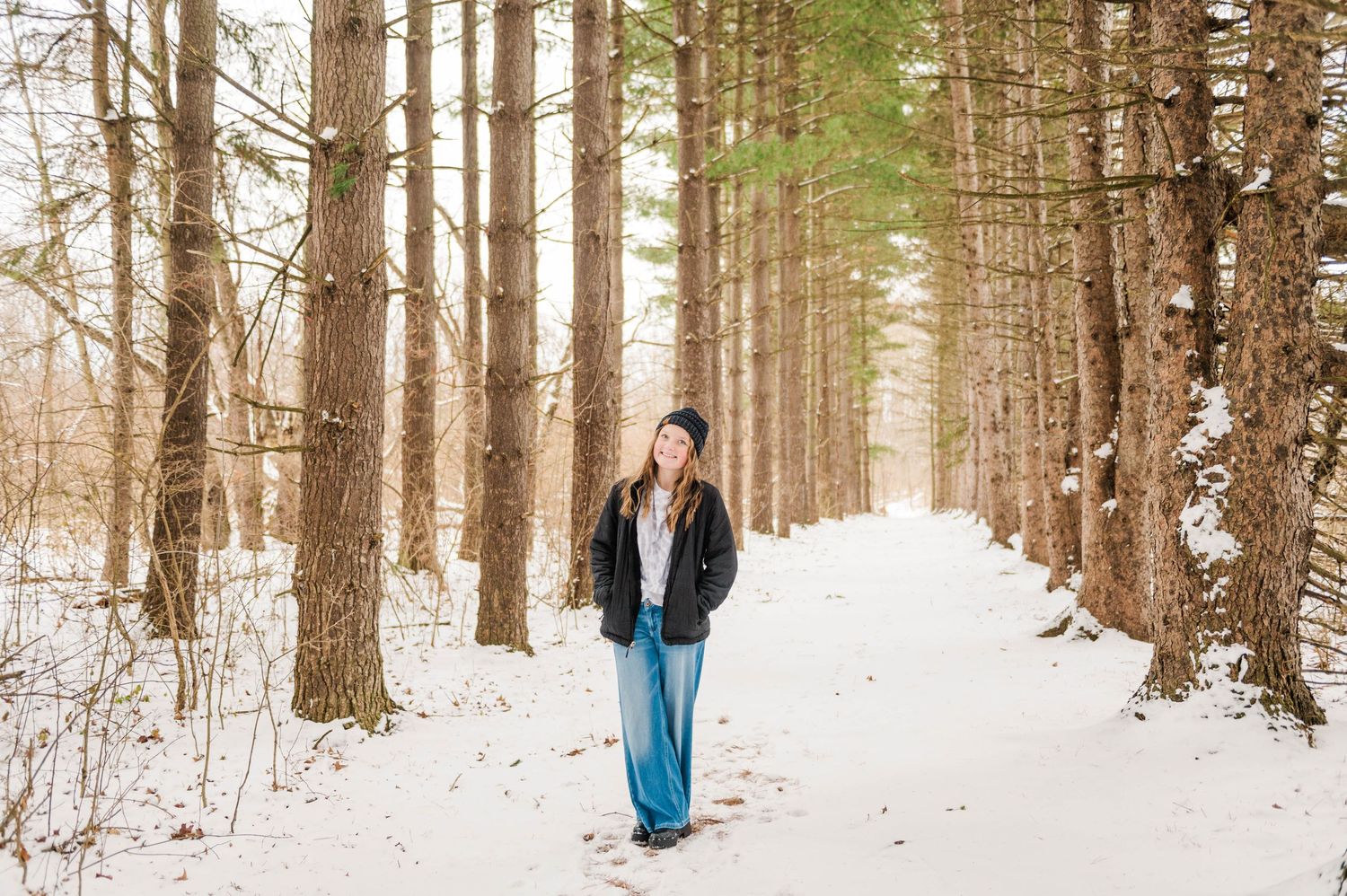 Snow-covered forest path lined with tall evergreen trees in winter.