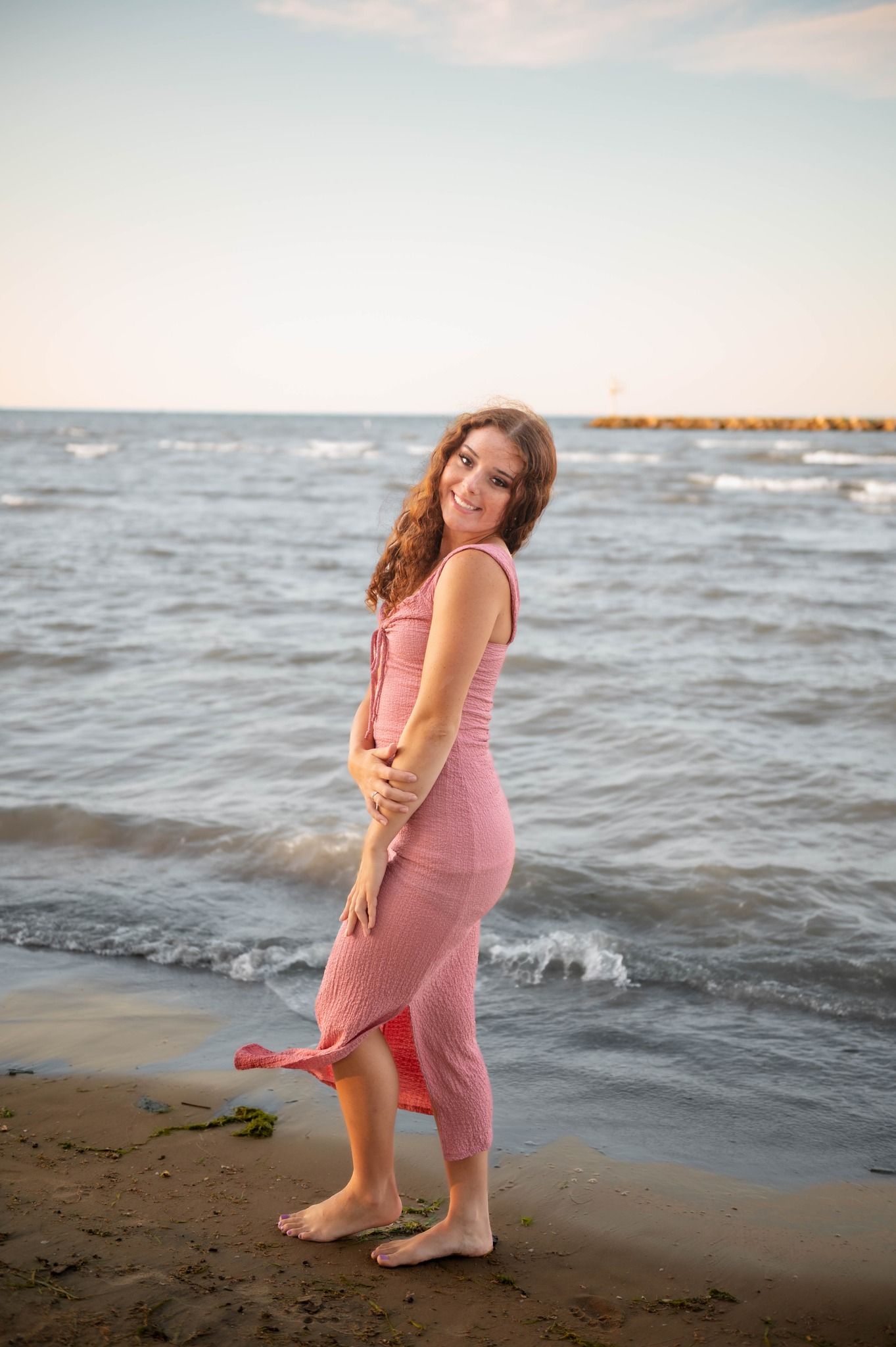 Someone in a pink dress stands playfully at the beach shoreline during sunset.