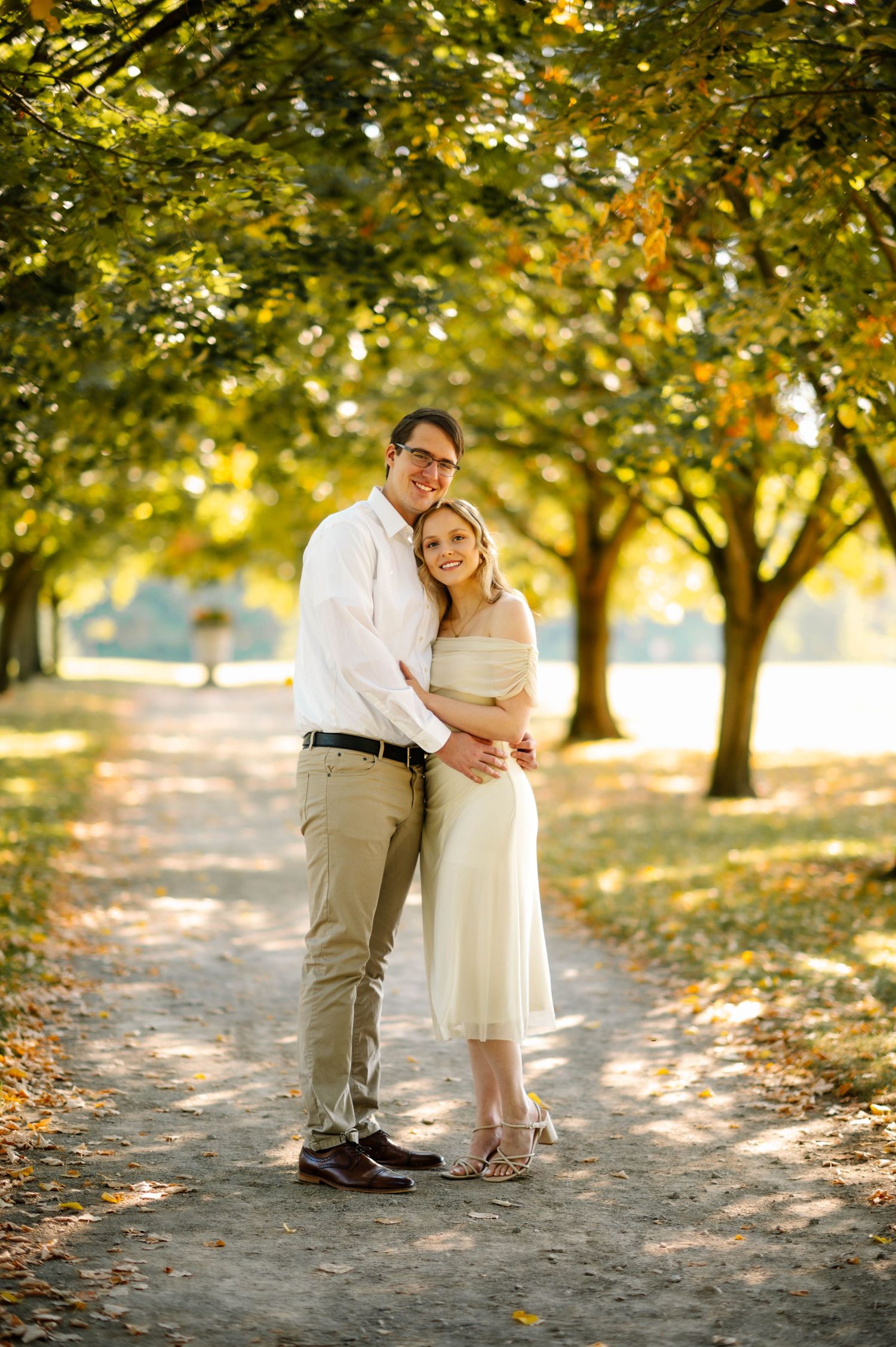 A couple embraces on a leaf-covered path beneath golden autumn trees.