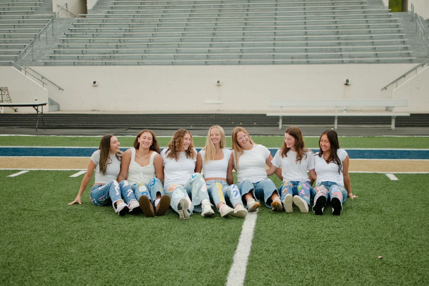 Seven girls in white tops and jeans sitting together laughing on a football field turf.