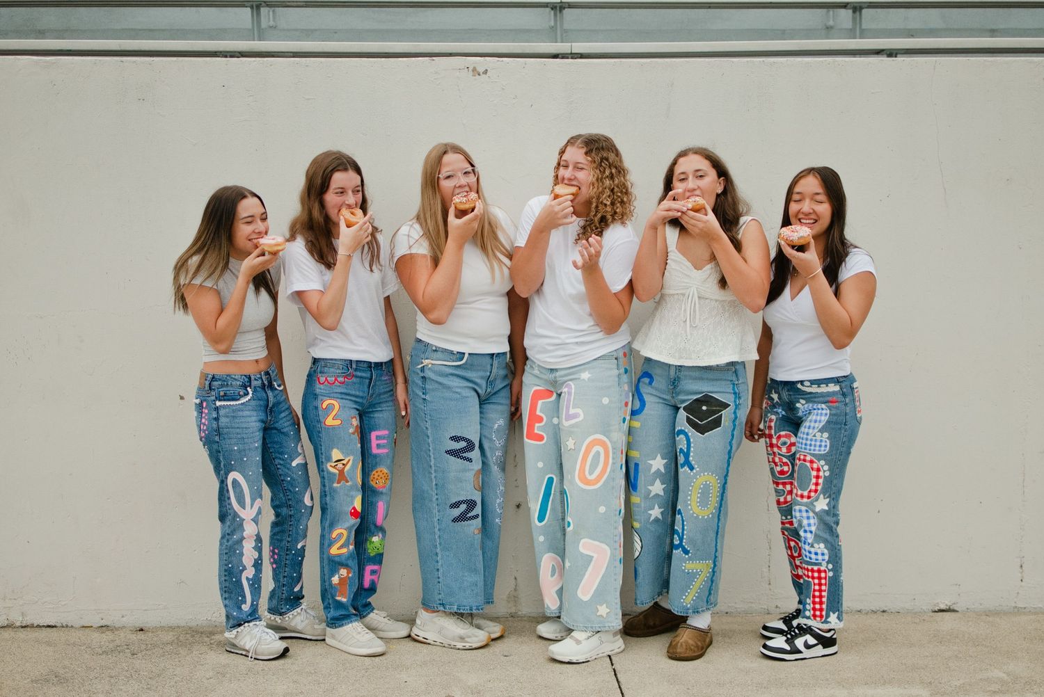 Six smiling girls in decorated jeans and white tops eating donuts against a wall.