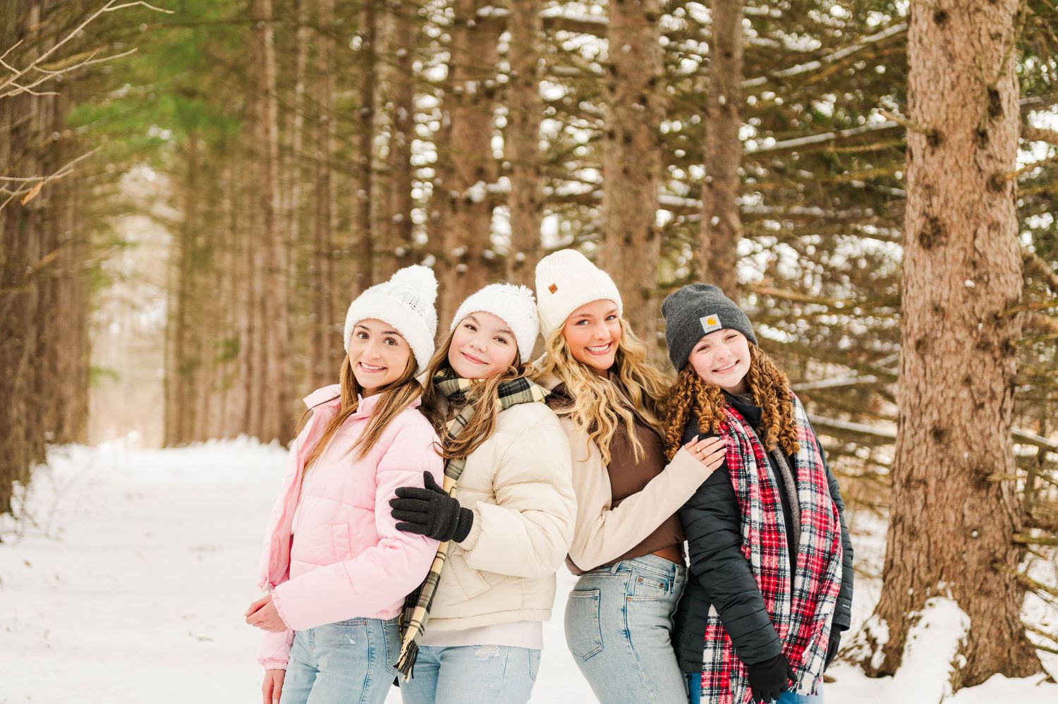 Group of friends wearing winter attire smile and pose back-to-back on a snow-covered forest trail.