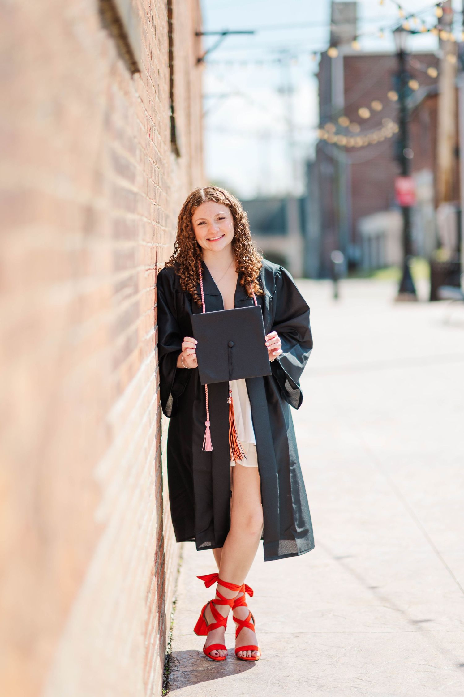 Curly-haired graduate in black gown and red heels holding cap against sunny brick wall.