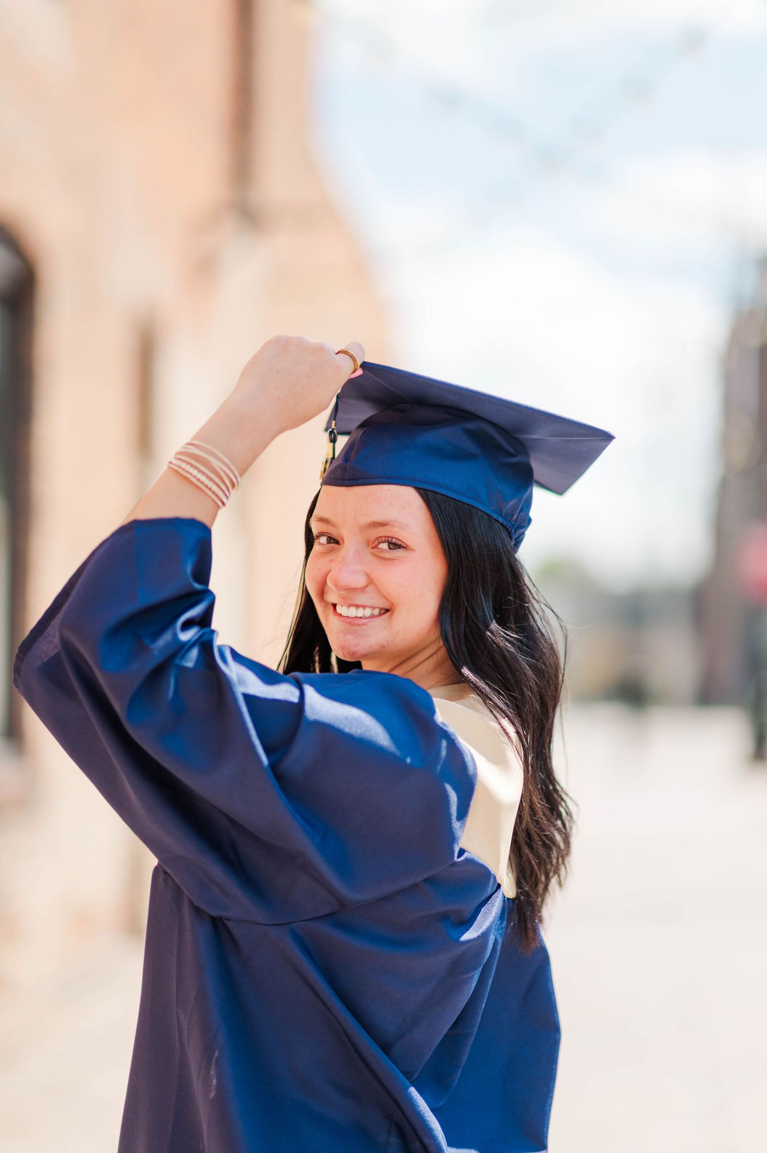 Smiling brunette woman in navy graduation cap and gown posing on sunny urban street.