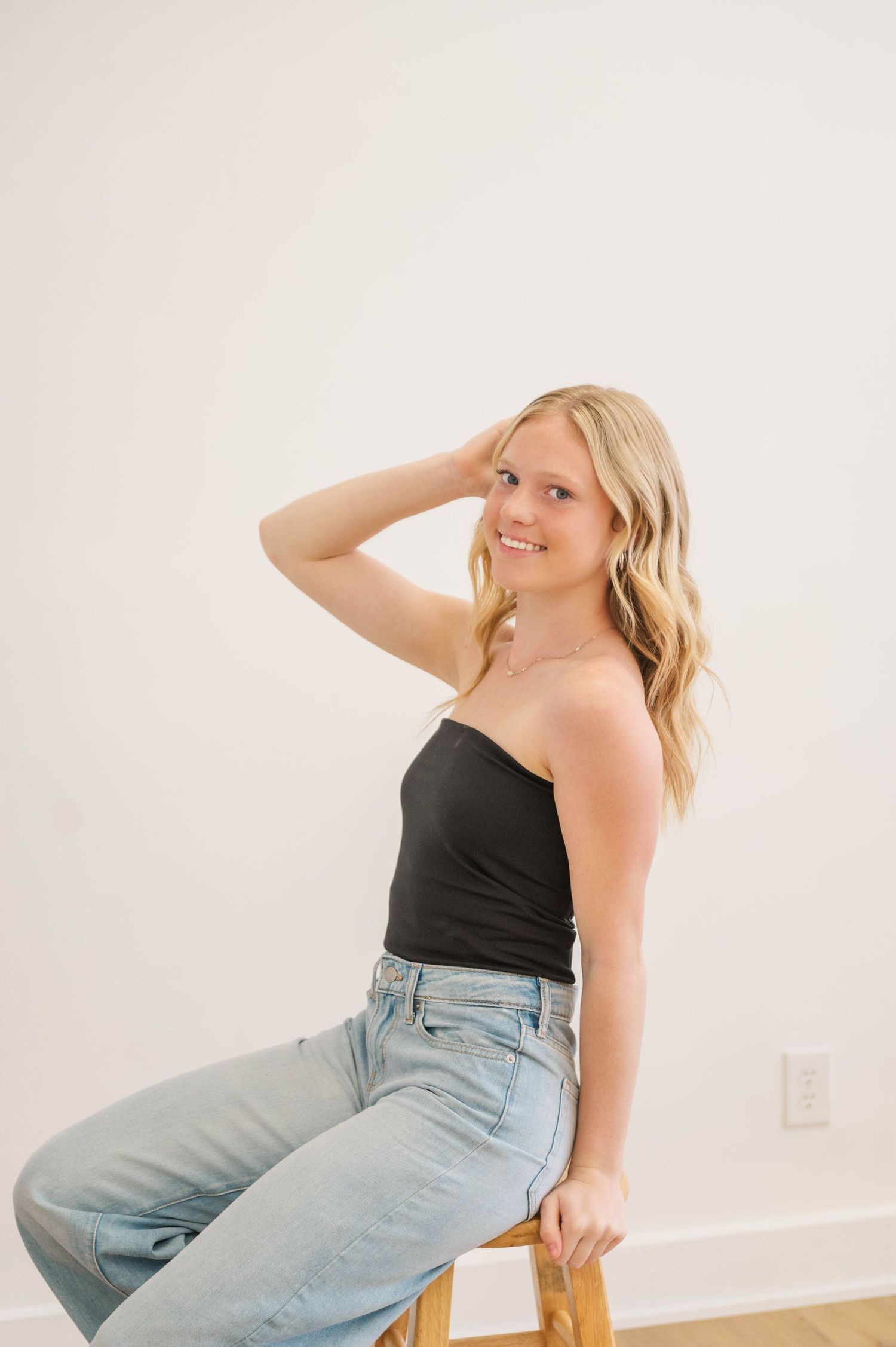 Blonde woman sits on wooden stool smiling, wearing black strapless top and light wash jeans, hand in hair.