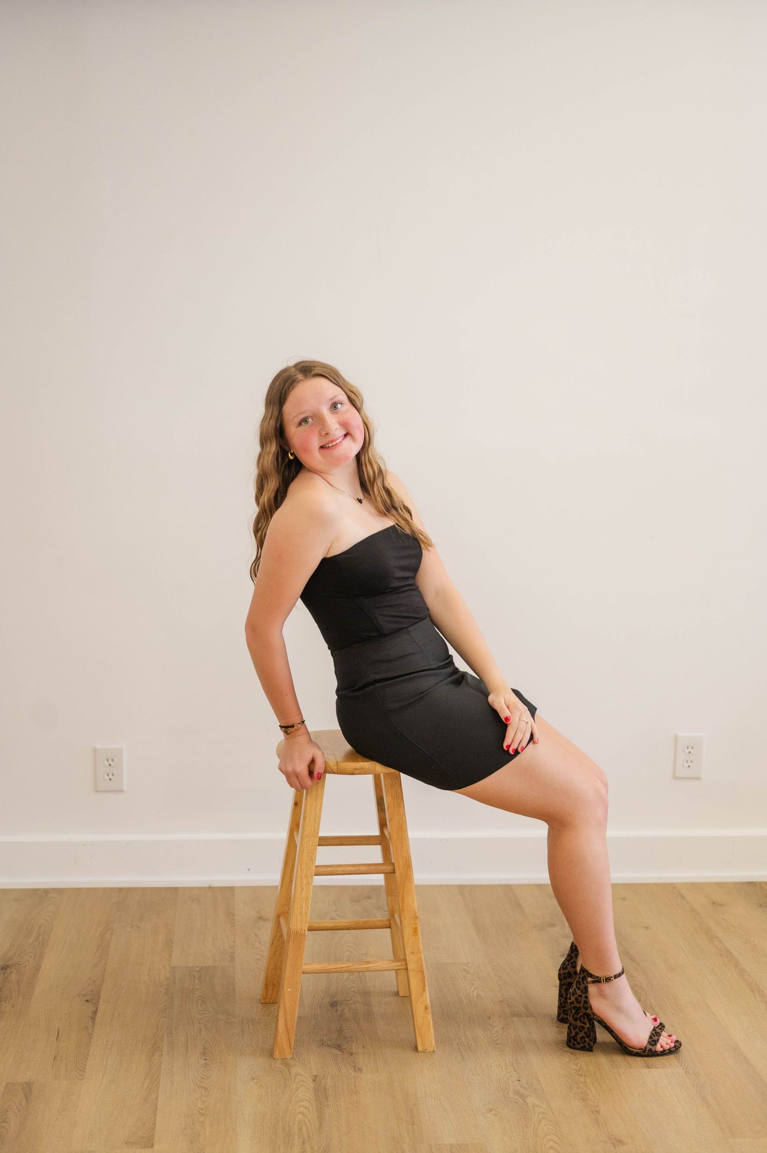 Young woman in black strapless mini dress sits on wooden stool wearing leopard print heels, smiling.