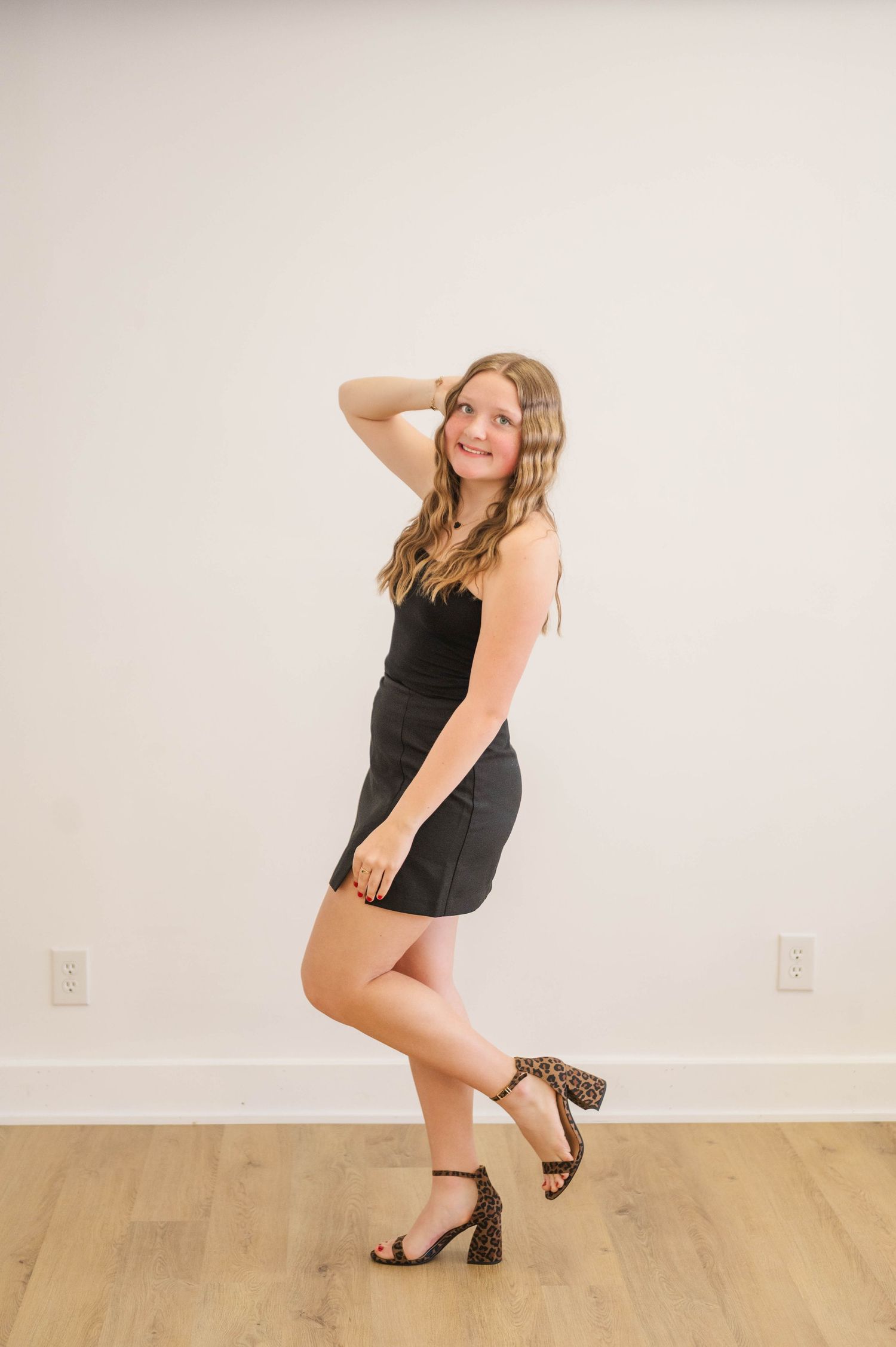 Blonde teen in black mini dress posing with hand in hair wearing leopard block heels.