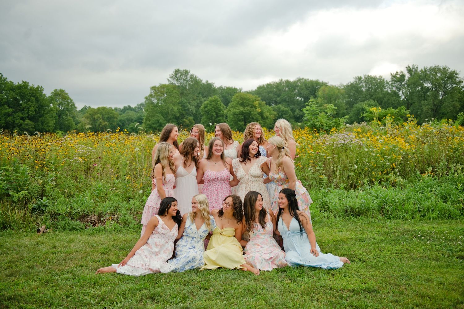 Group of girls in pastel dresses posing together in a lush green field with yellow wildflowers under cloudy skies.
