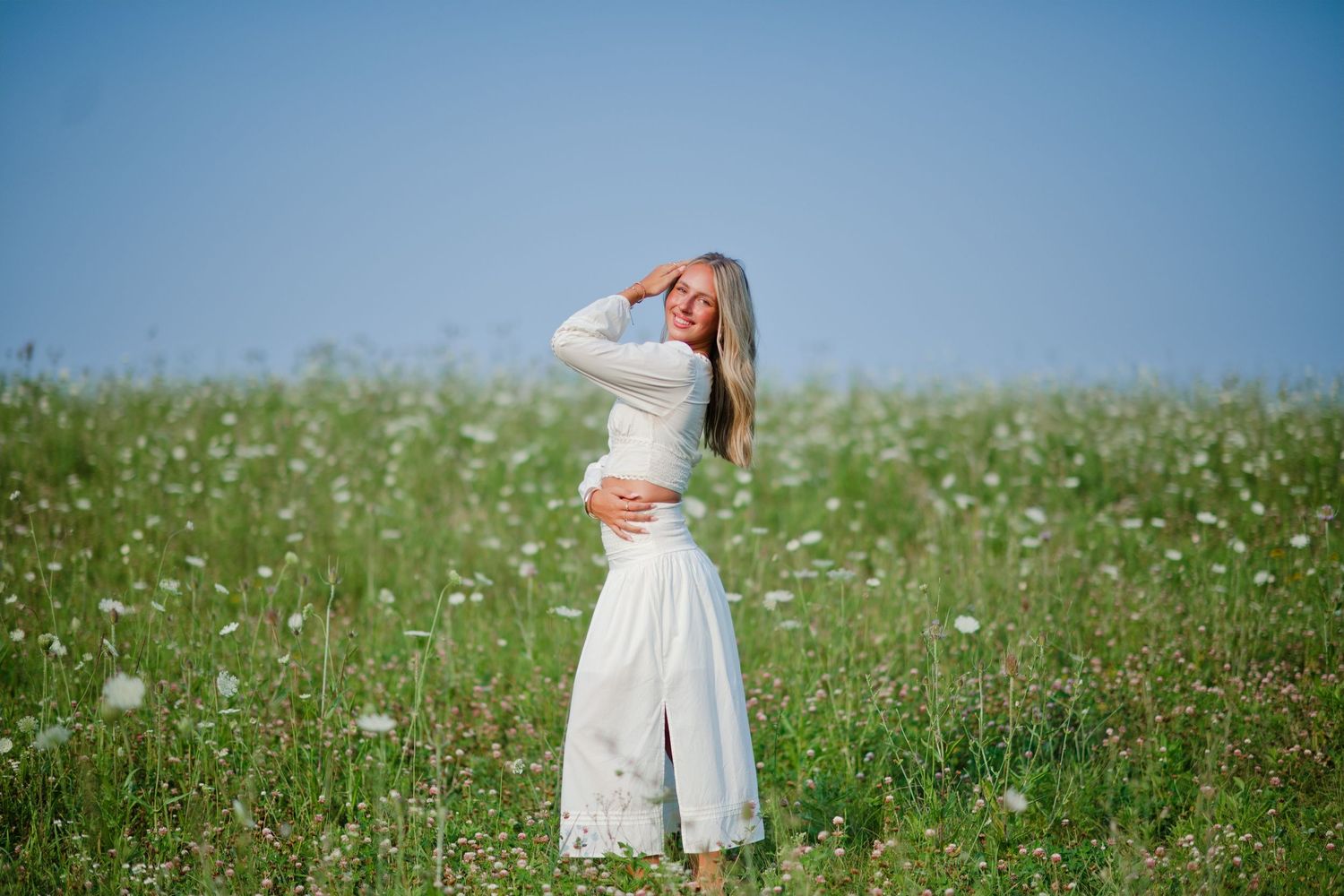 Woman in white dress standing in a blooming meadow under a clear blue sky, shielding her eyes from the sun.