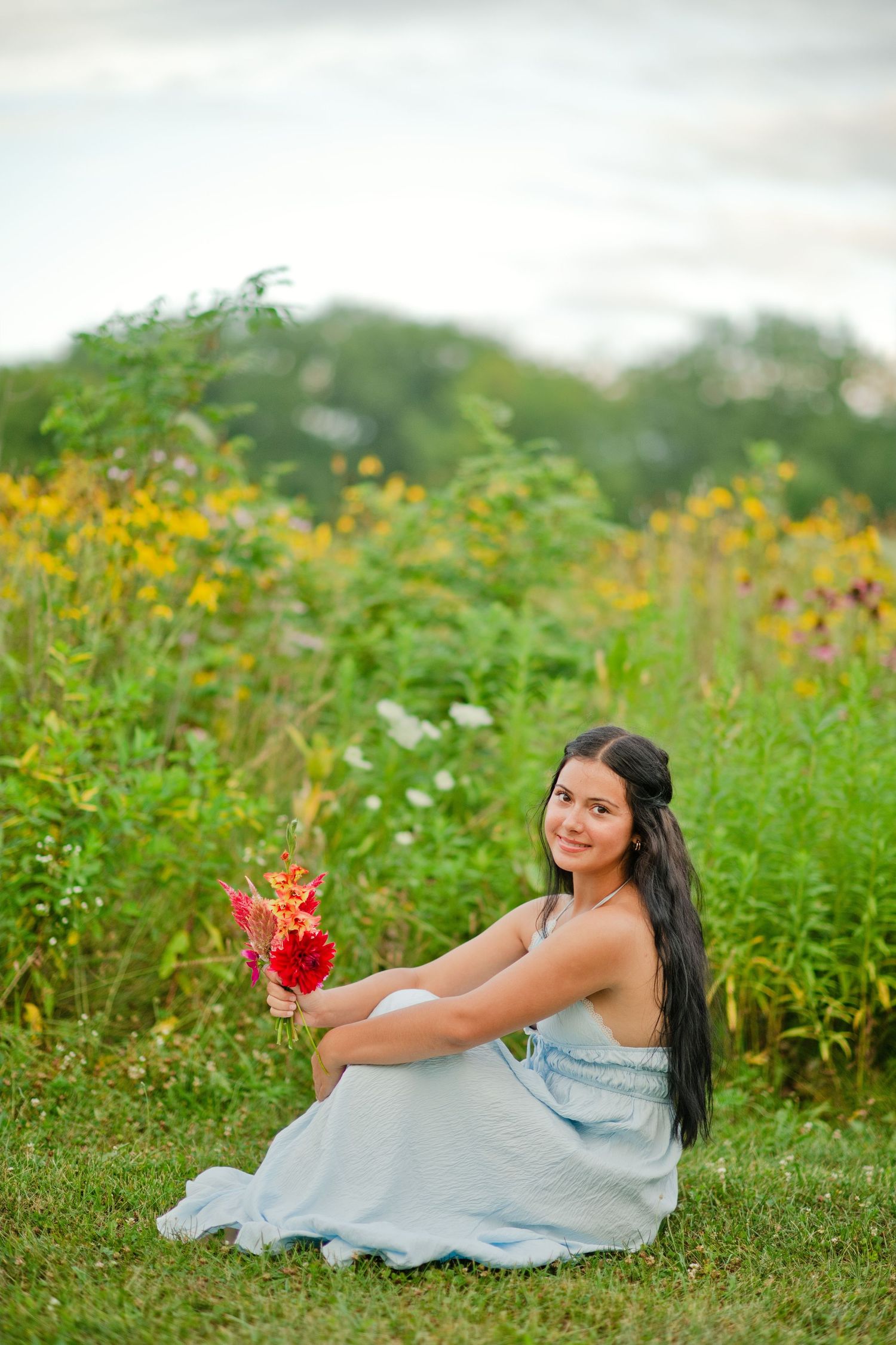 Young woman in light blue dress sitting in wildflower field holding red flowers, smiling outdoors.