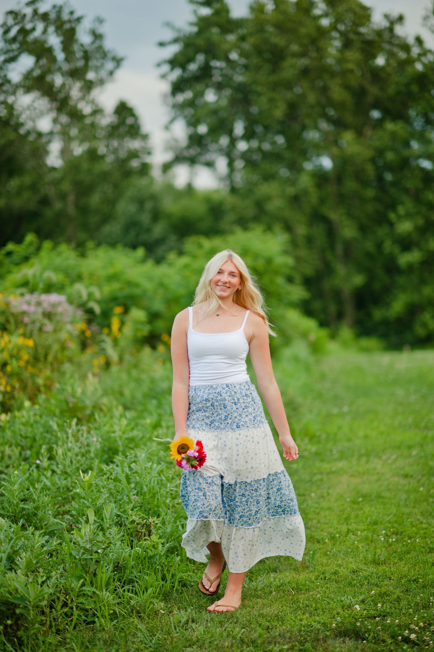 Smiling blonde woman in white tank top and blue floral maxi skirt walking barefoot on grass holding colorful bouquet.
