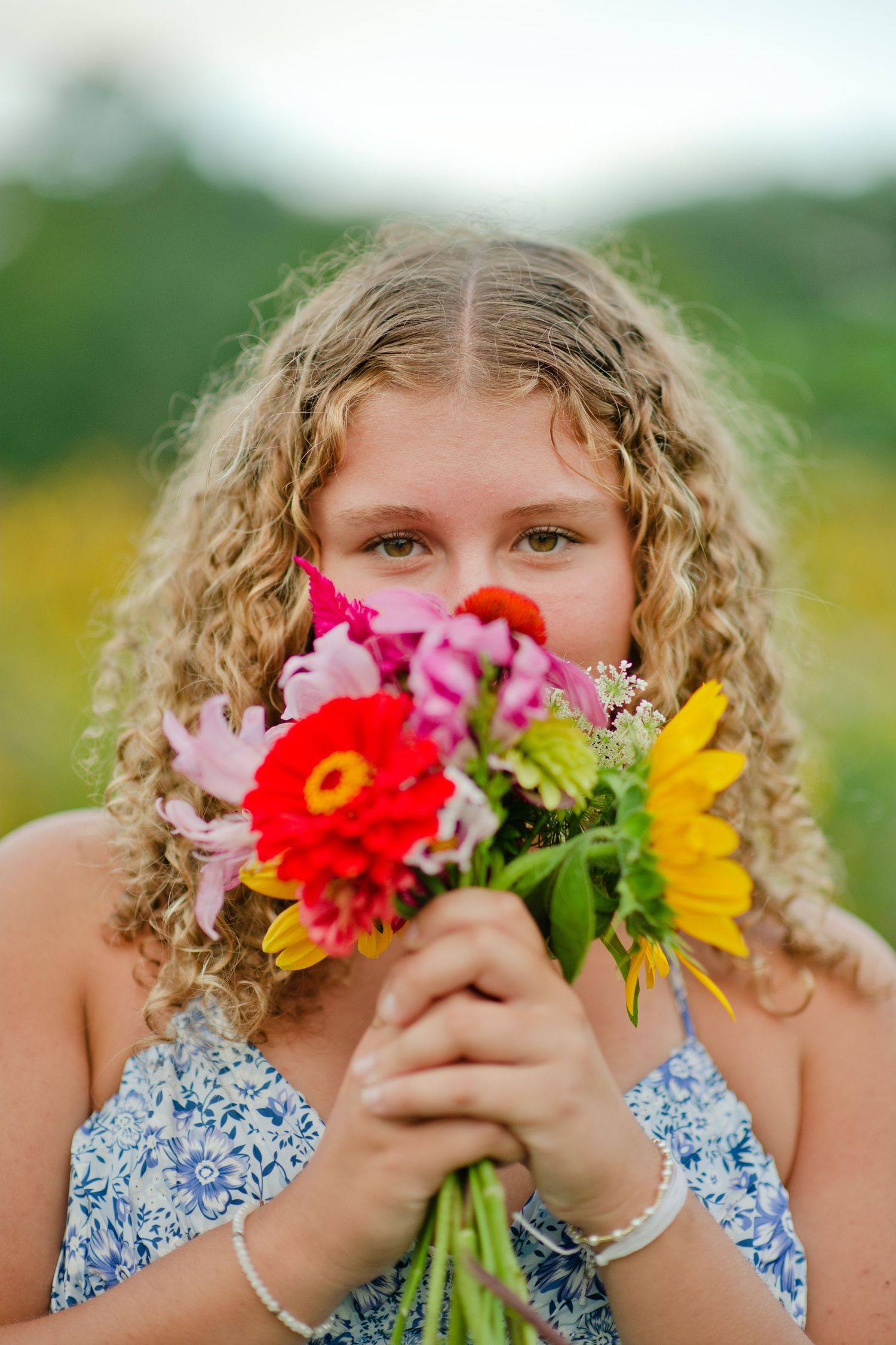 A curly-haired blonde girl holds a colorful bouquet of zinnias and sunflowers up to her face outdoors.