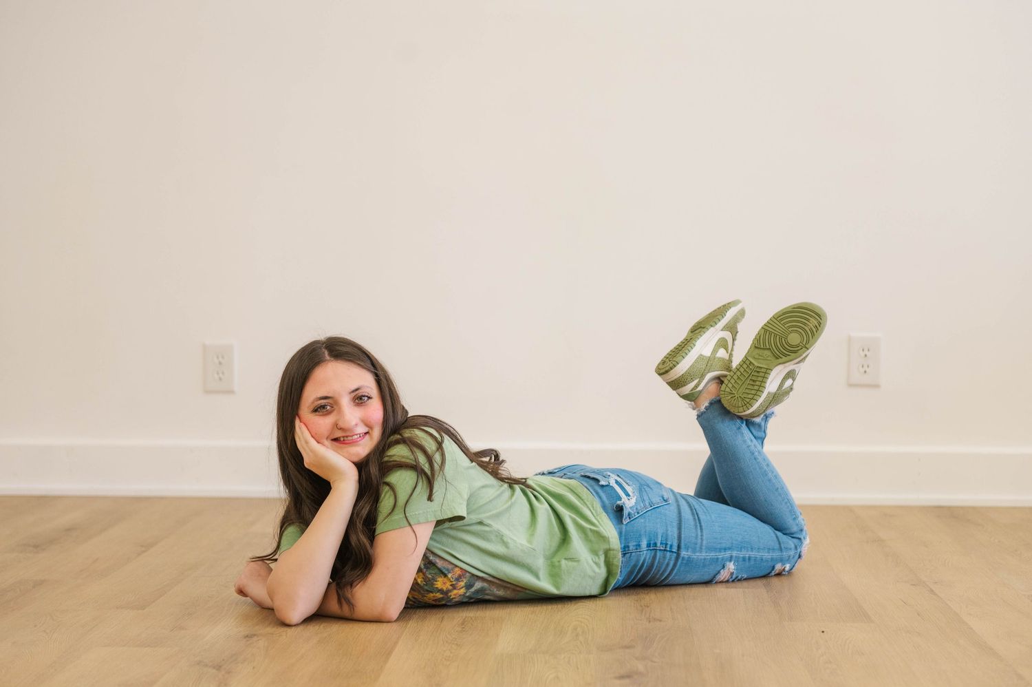 Brunette girl in green tee and ripped jeans lying on wood floor smiling at camera.