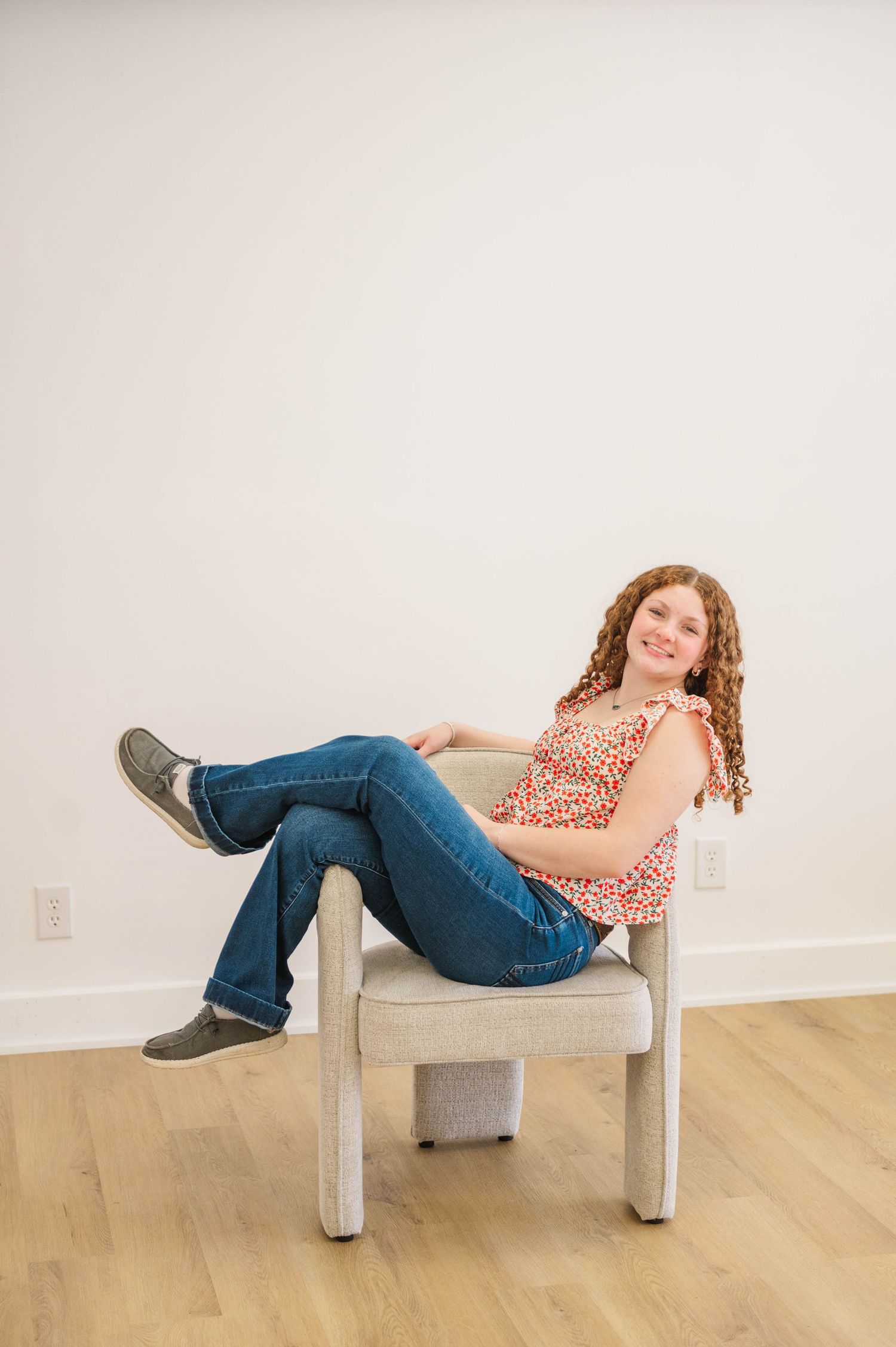 Curly-haired girl in floral top and jeans posing casually in a light gray accent chair.