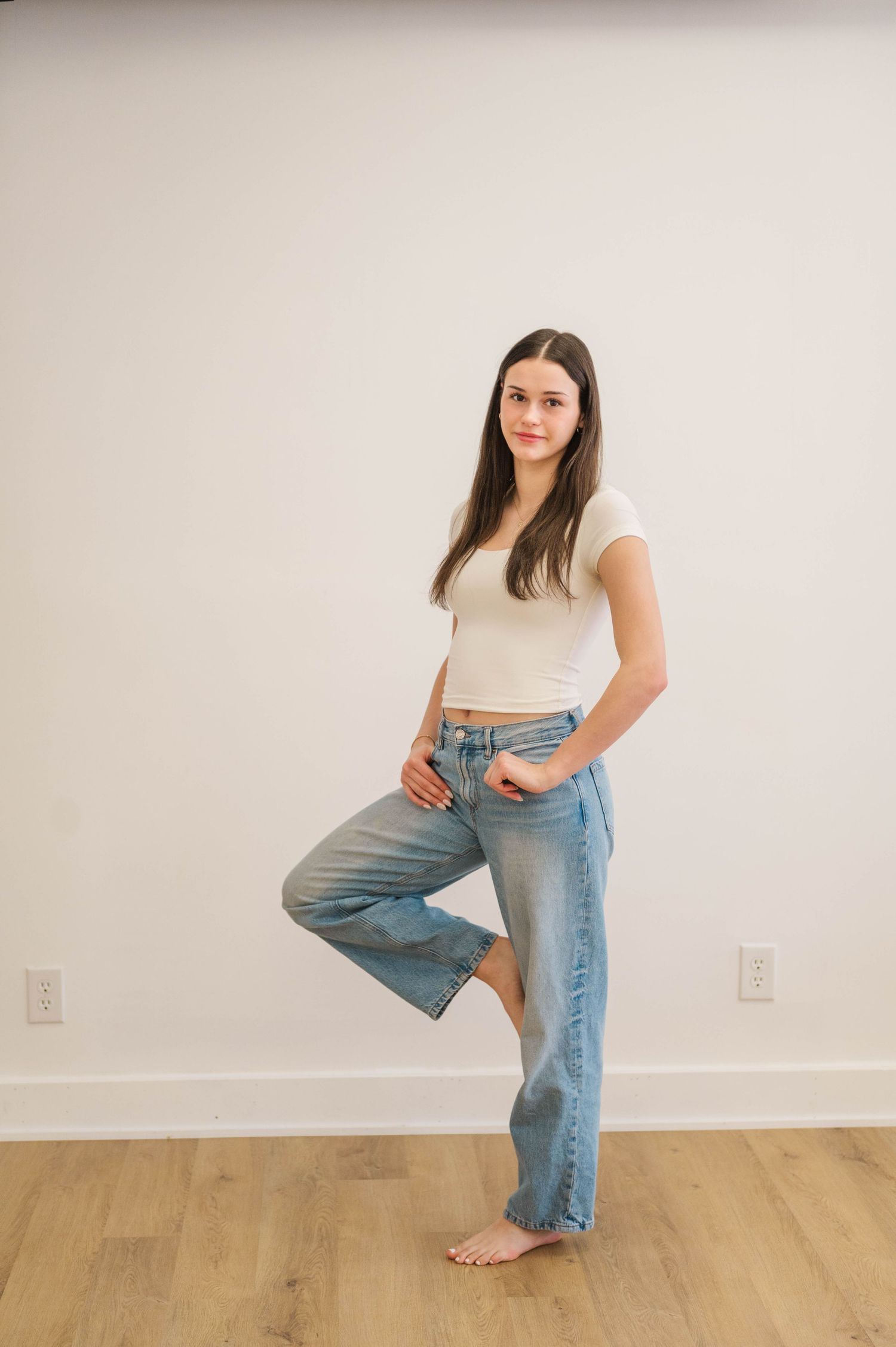 Young woman in white crop top and wide-leg jeans stands barefoot on one leg against white wall.