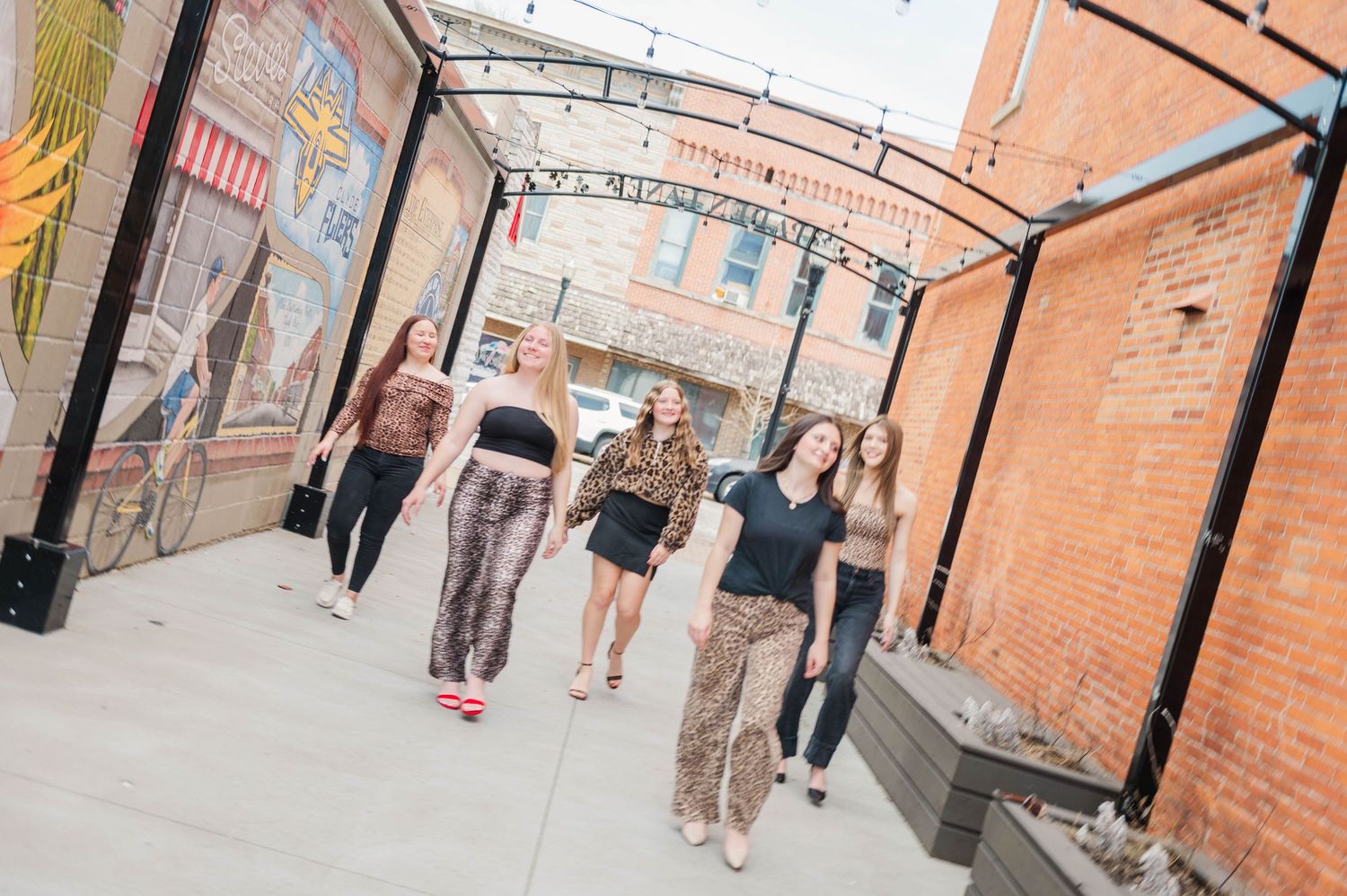 Five young women walking together in an urban alley with brick walls and colorful murals.