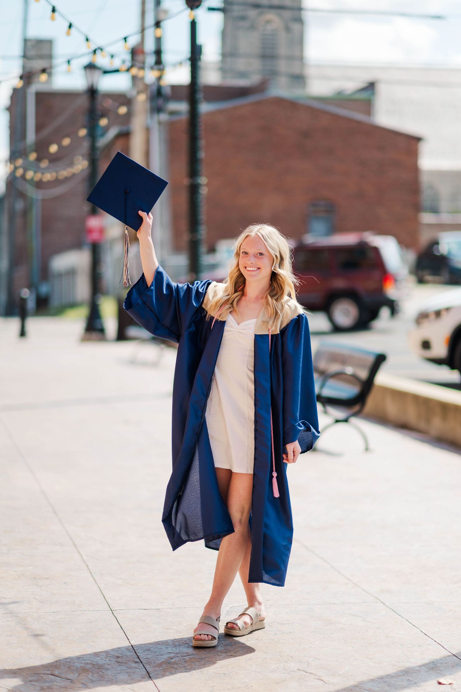 Smiling blonde graduate in navy gown holding cap in the air while walking on urban sidewalk.