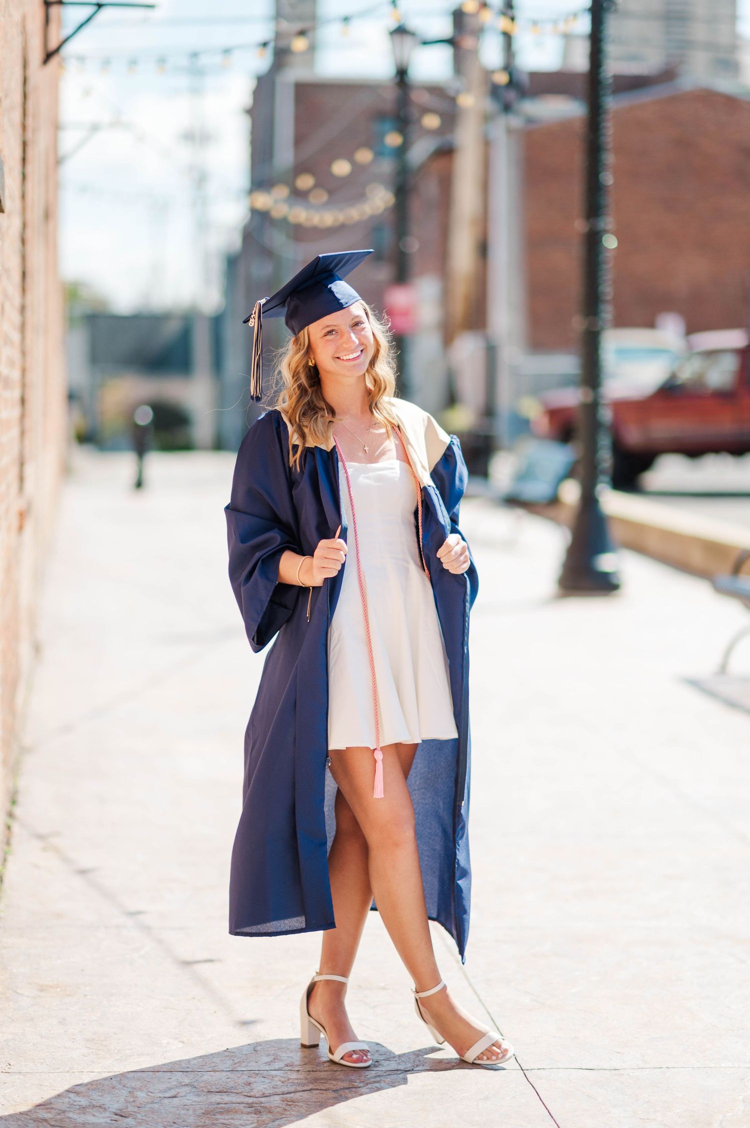 Smiling graduate in navy cap and gown walking on sunny urban sidewalk wearing white dress underneath.