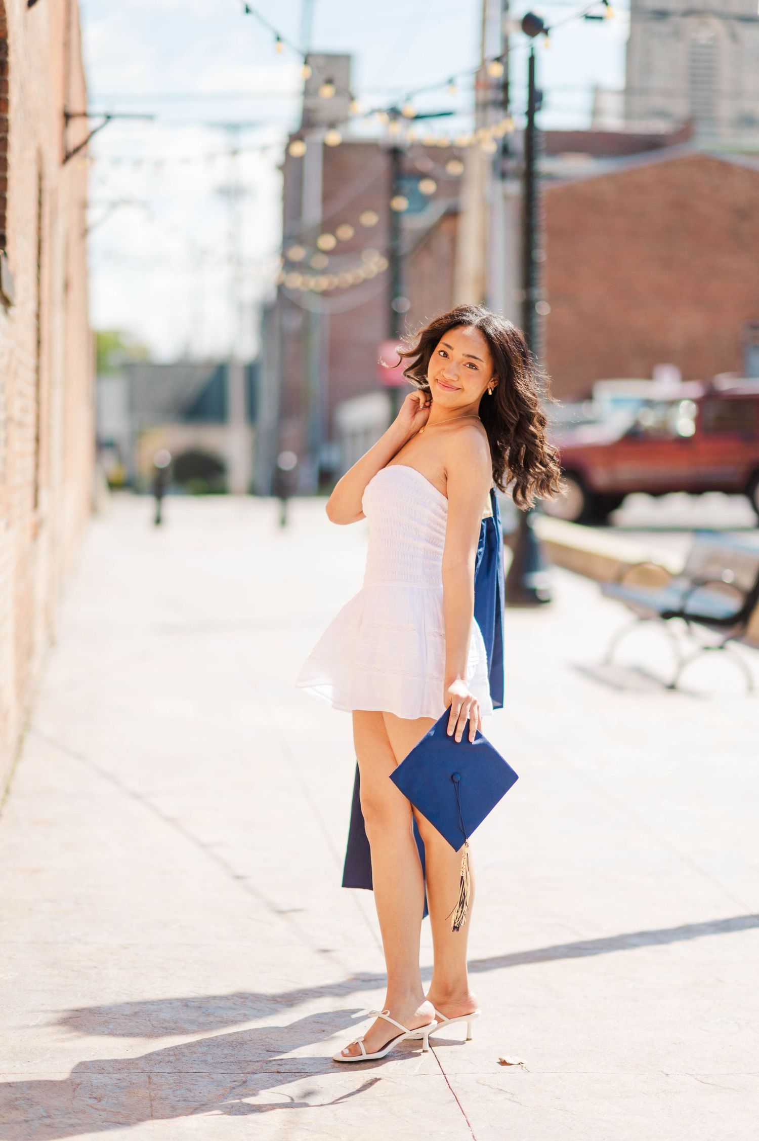 Young woman in white strapless dress holding blue graduation cap and gown outdoors on sunny brick sidewalk.