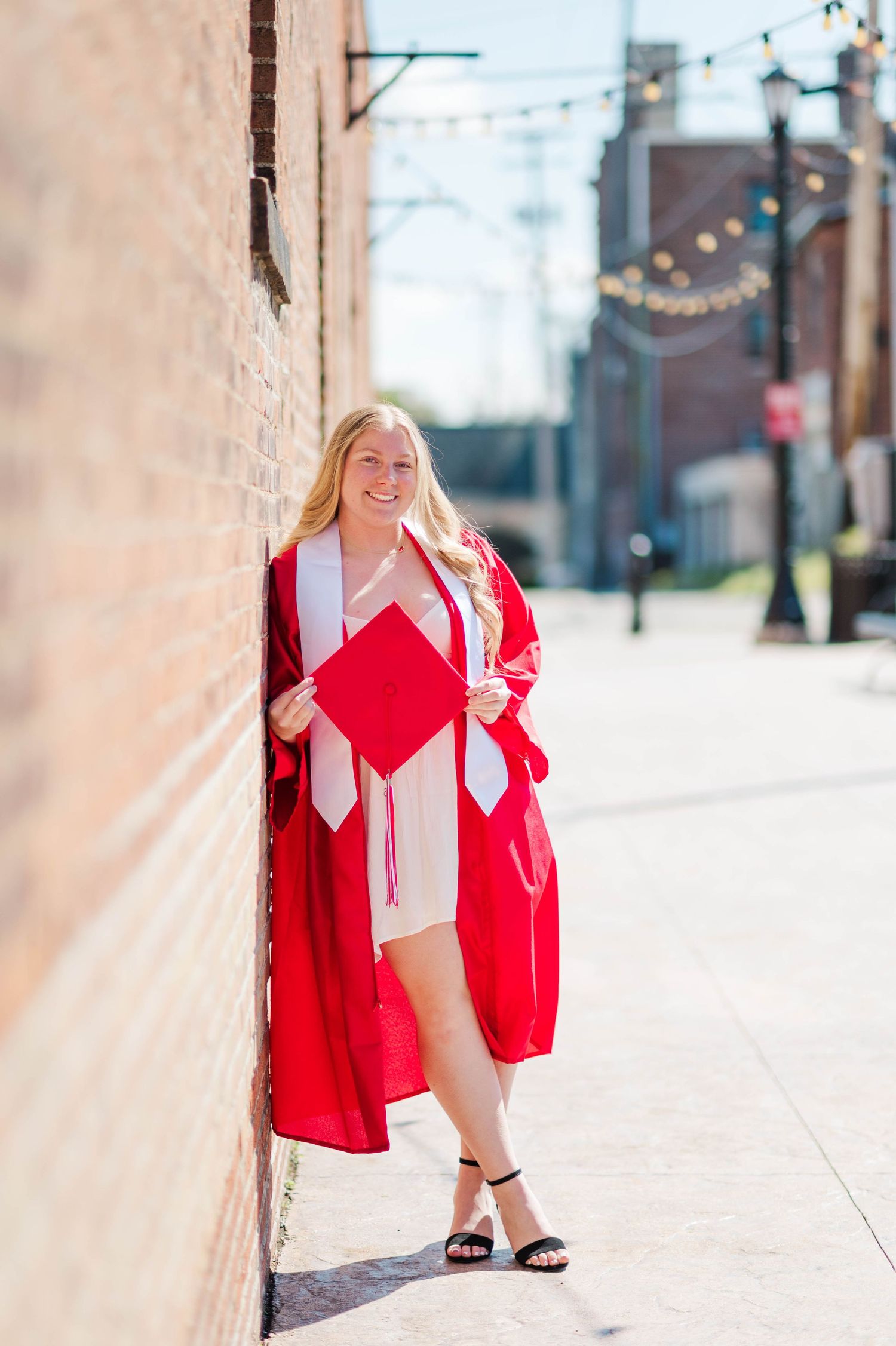 Happy graduate in red cap and gown leaning against brick wall smiling on sunny urban sidewalk.