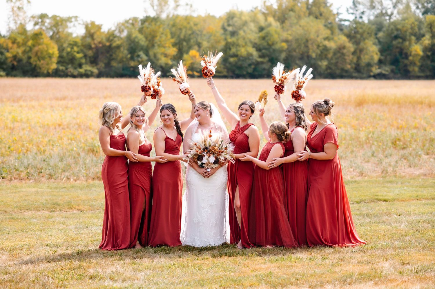 Bridal party in flowing red dresses poses together in sunny autumn field.