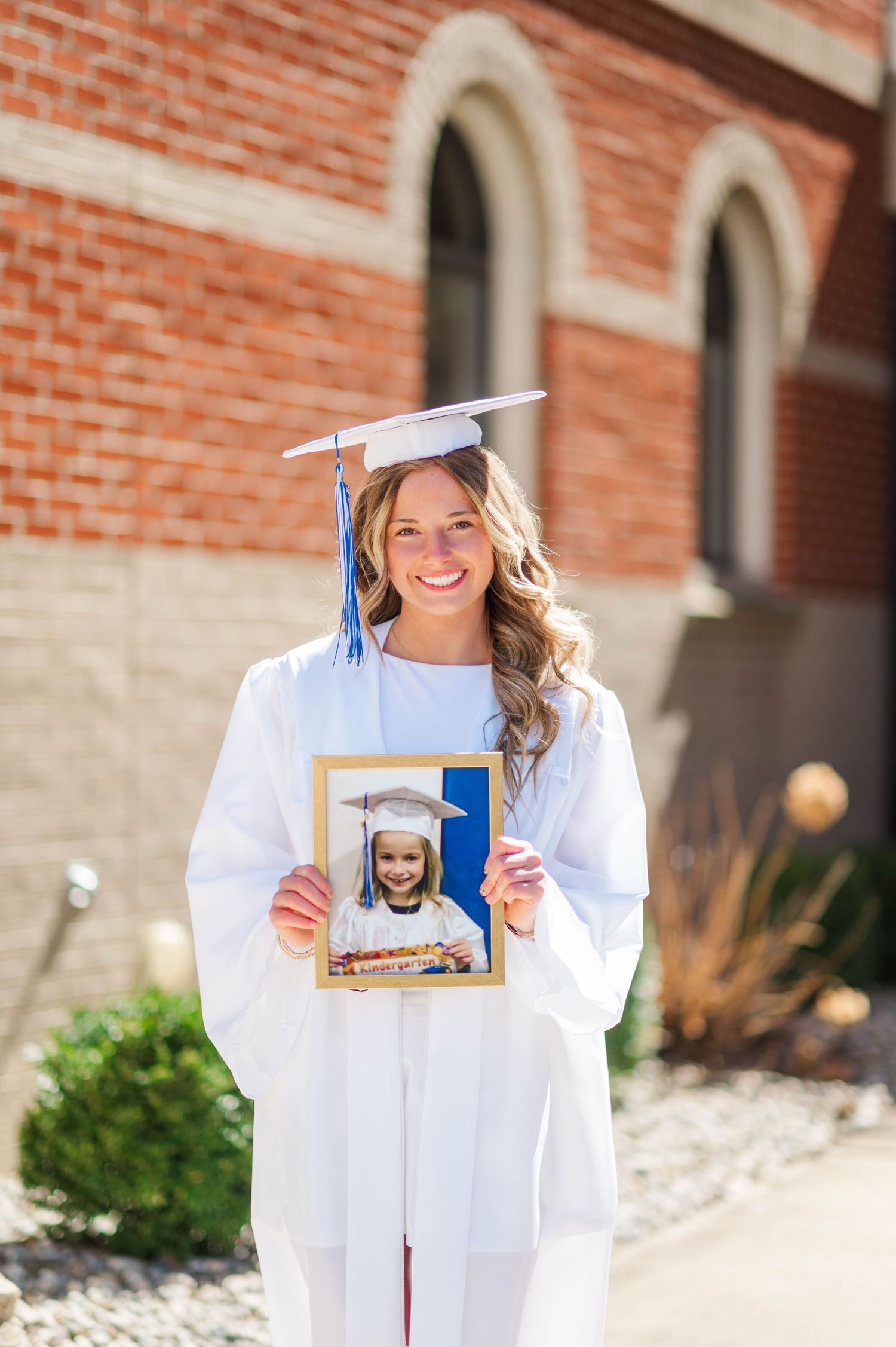 Graduate in white cap and gown holding framed photo during outdoor celebration shoot.