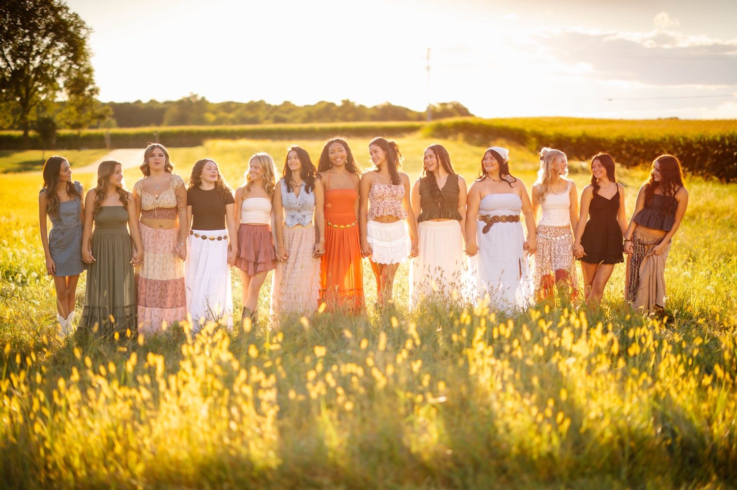 A line of people in colorful dresses pose together in a sunlit meadow.