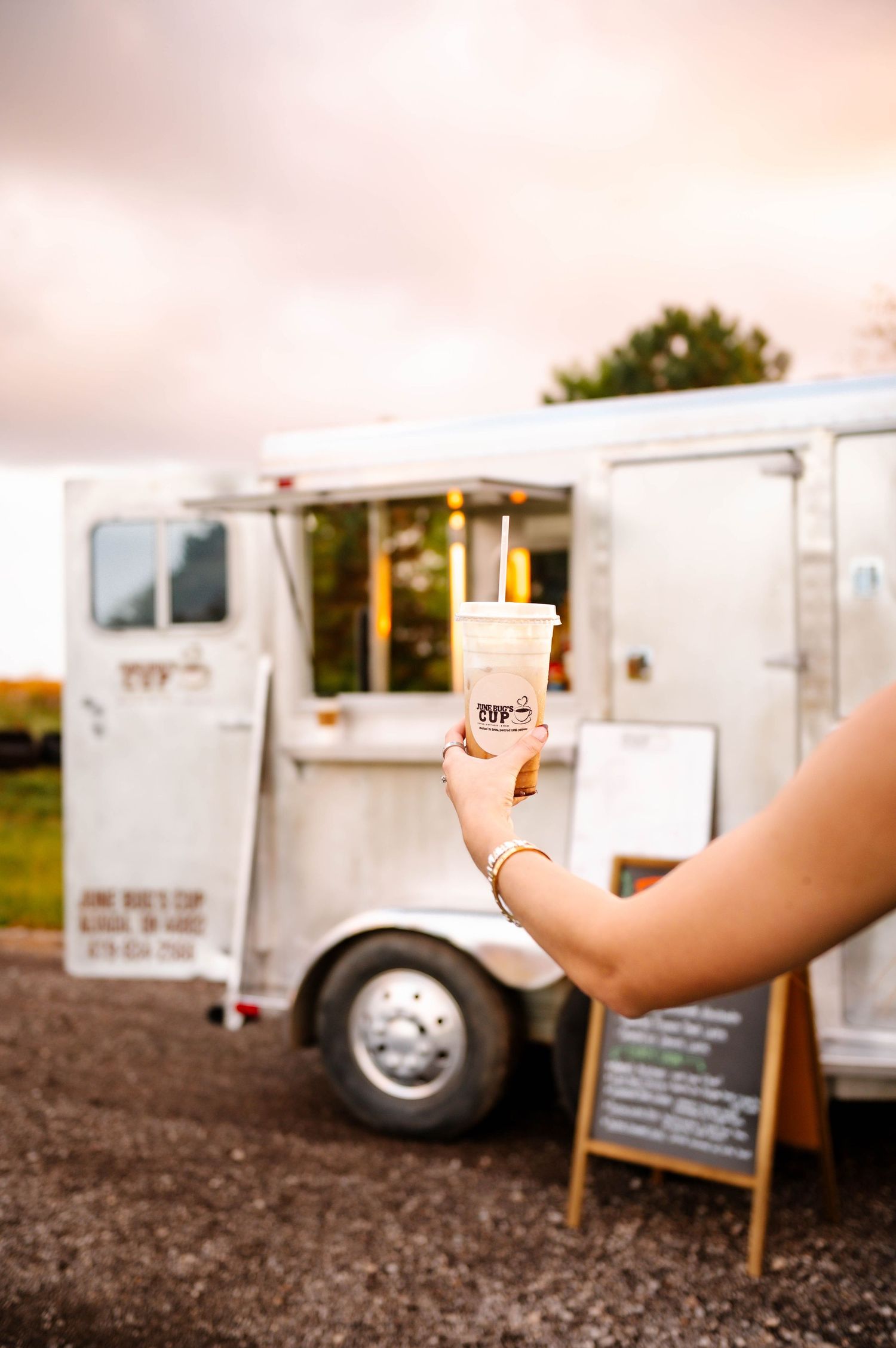 Arm holding coffee cup against backdrop of white food trucks at sunset.