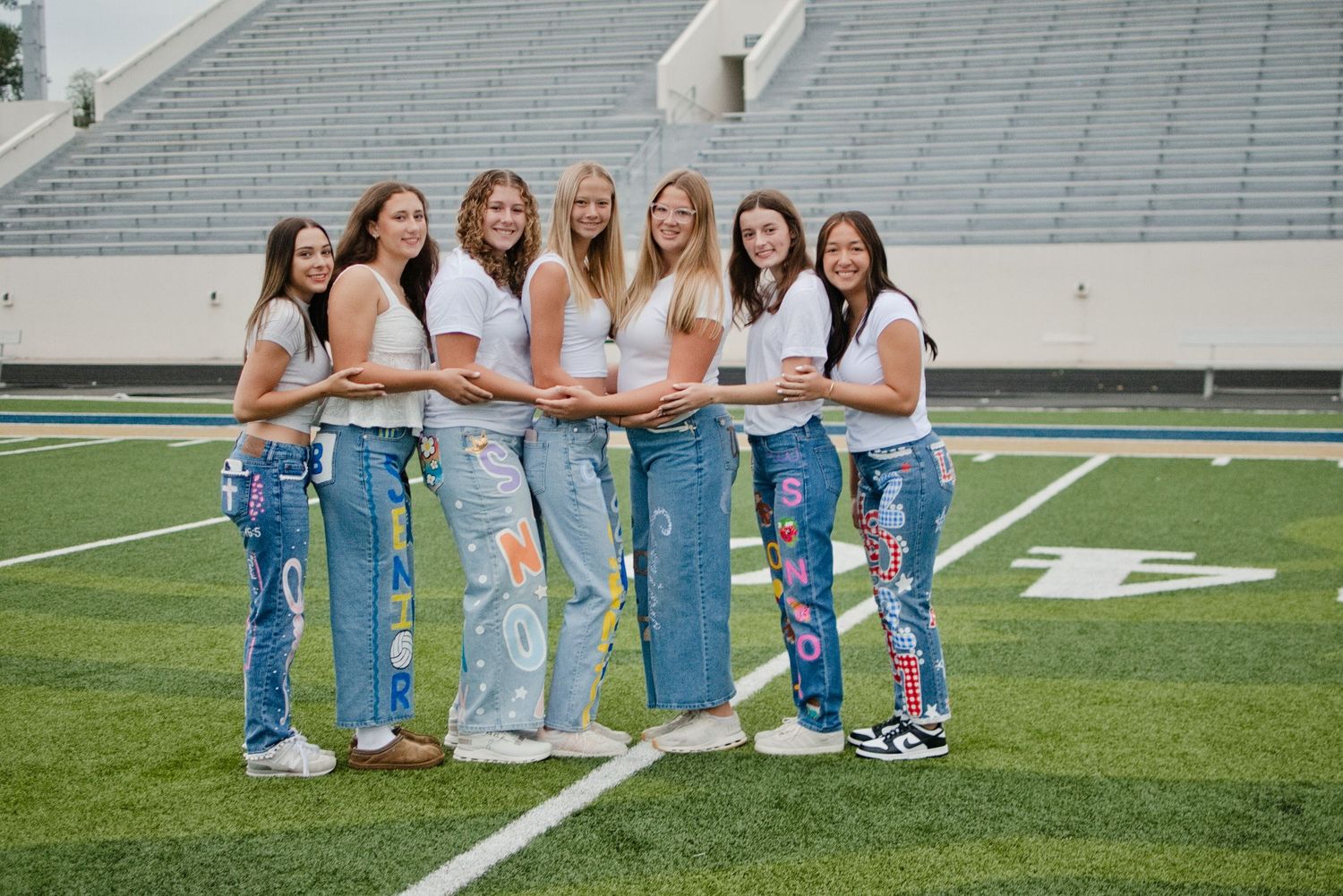 Six young women pose together on a football field wearing white tops and decorated denim jeans.