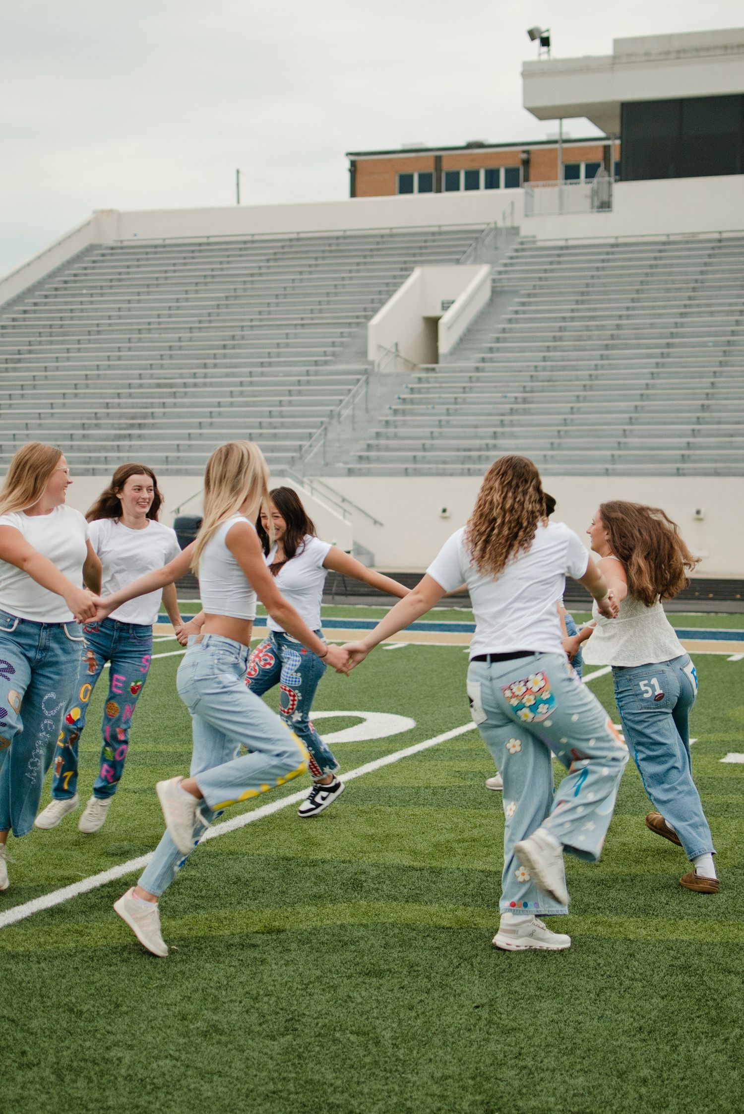 Girls in white tees and painted jeans hold hands dancing on a football field stadium in background.
