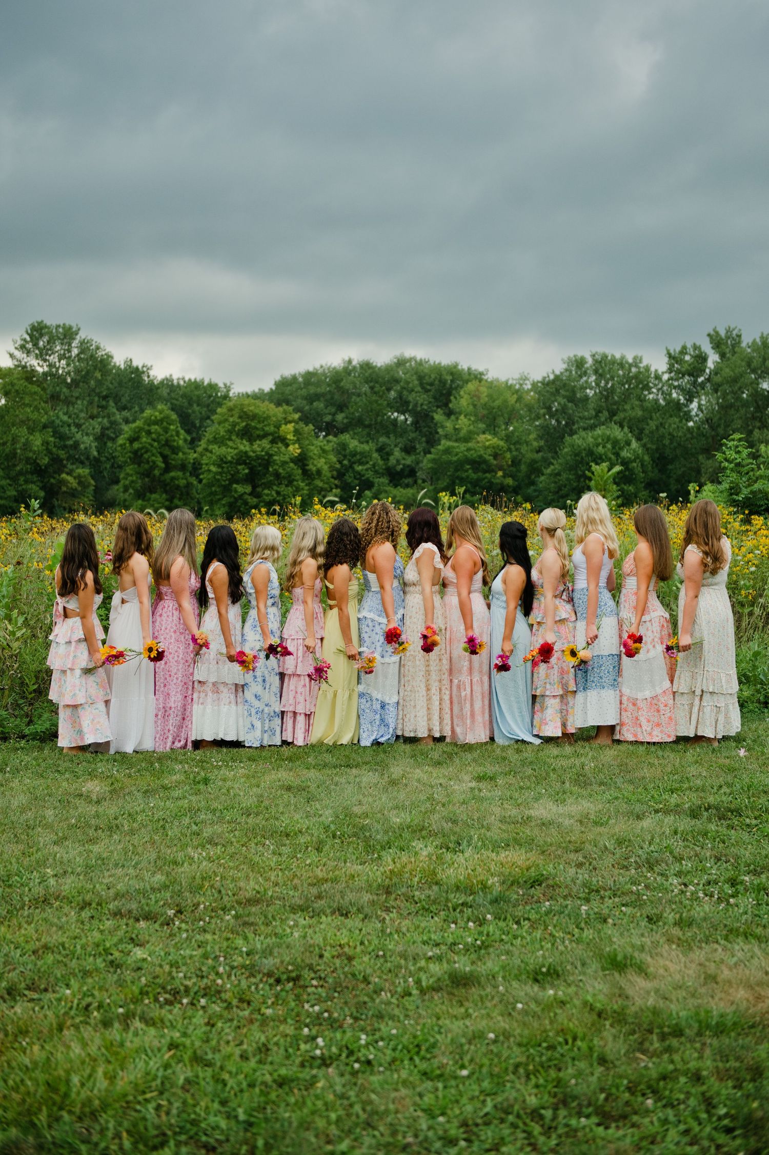 A group of bridesmaids in colorful mismatched dresses stand together facing a wildflower field on a cloudy day.