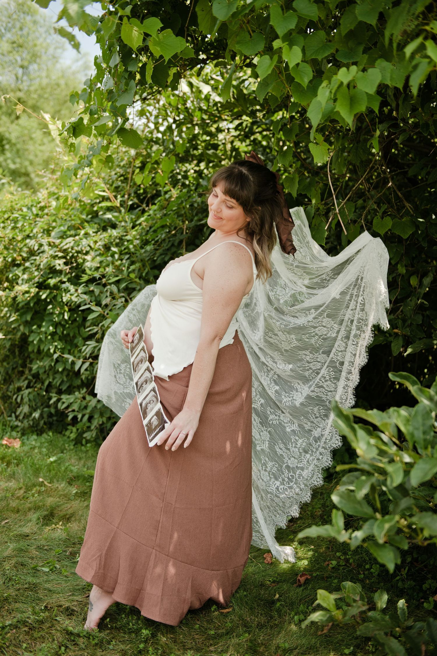 Pregnant woman in white lace veil and mauve skirt holds ultrasound photo outdoors among lush green foliage.