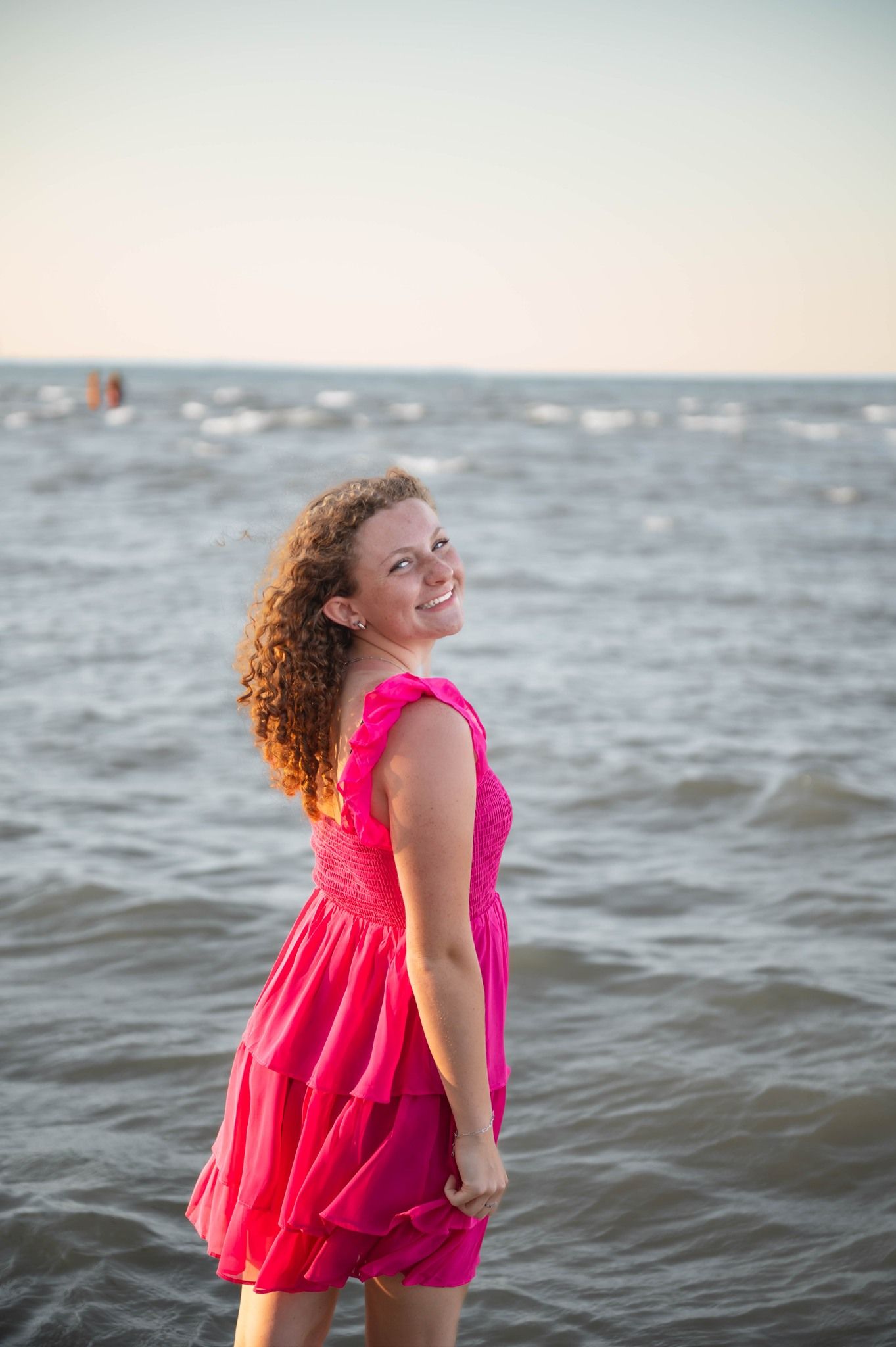 A person in a bright coral dress twirls joyfully by the ocean waves at sunset.