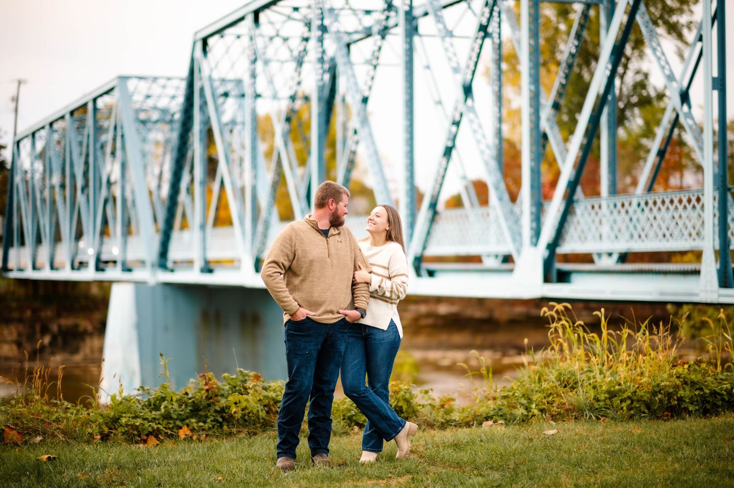 Couple walking hand-in-hand near a blue steel bridge surrounded by autumn foliage in a park setting.