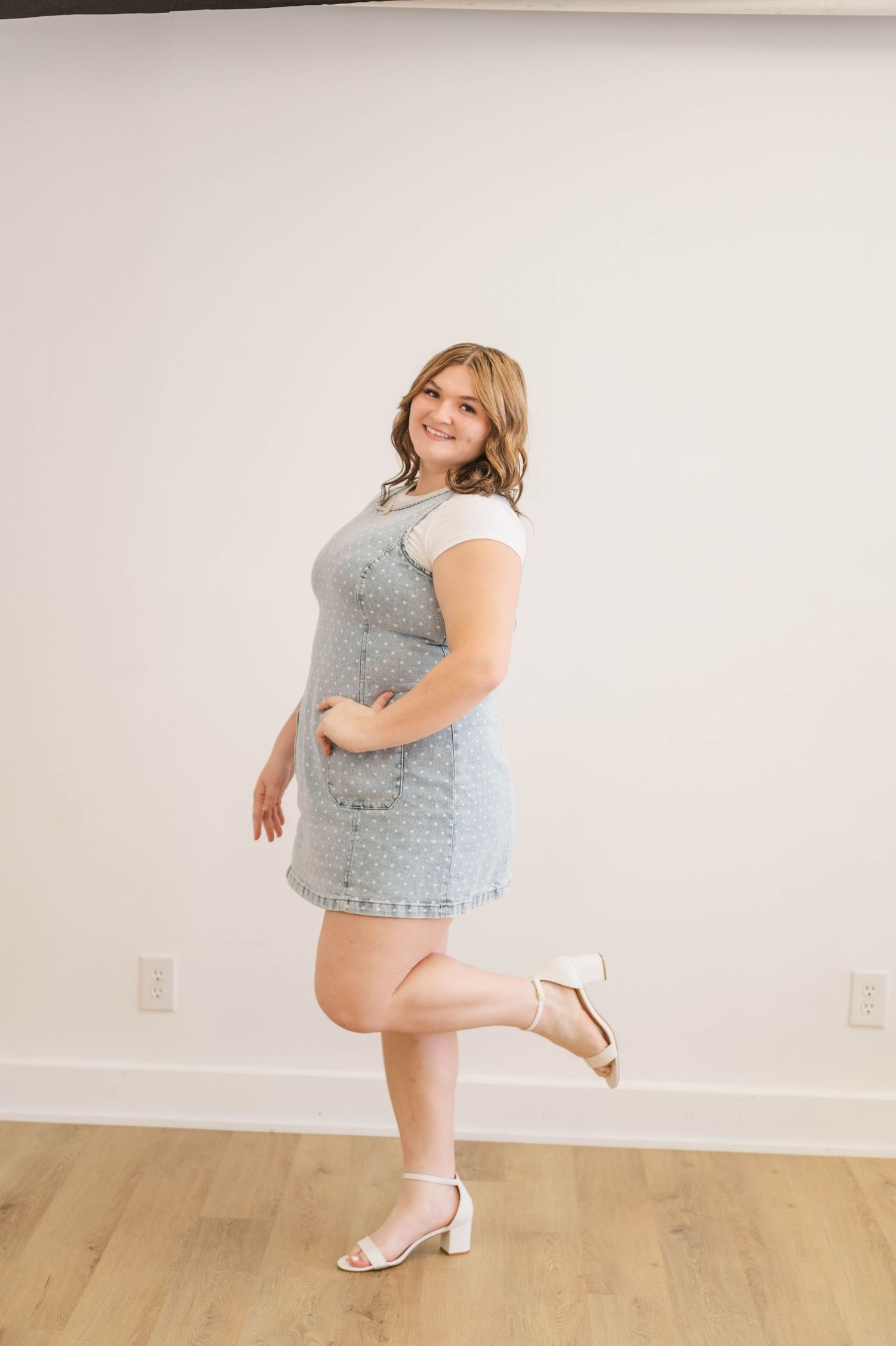 Smiling woman in light blue denim dress and white heels posing on one leg in bright studio.