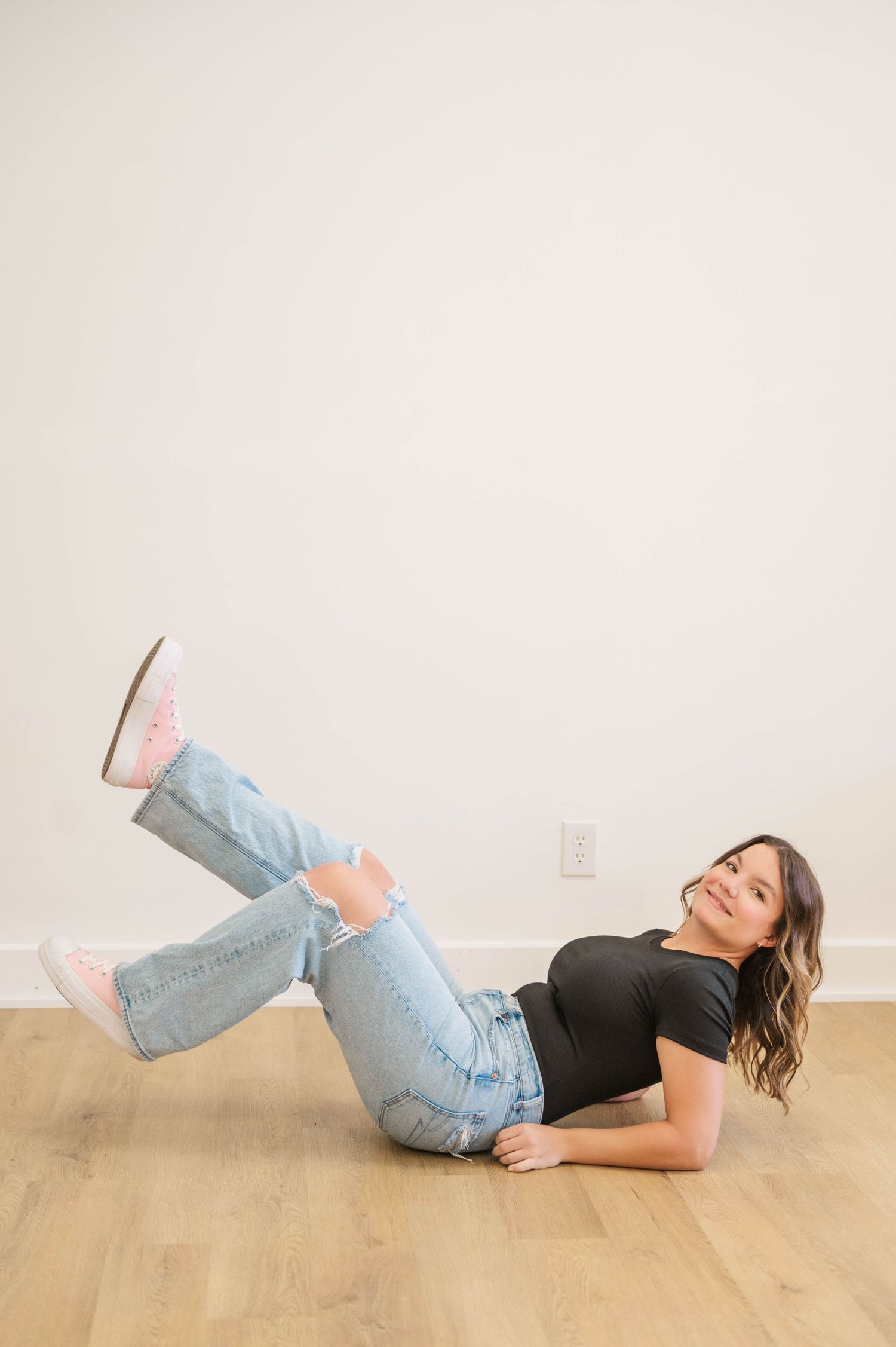 Young woman in black tee and ripped jeans lies on floor with legs raised, smiling playfully.