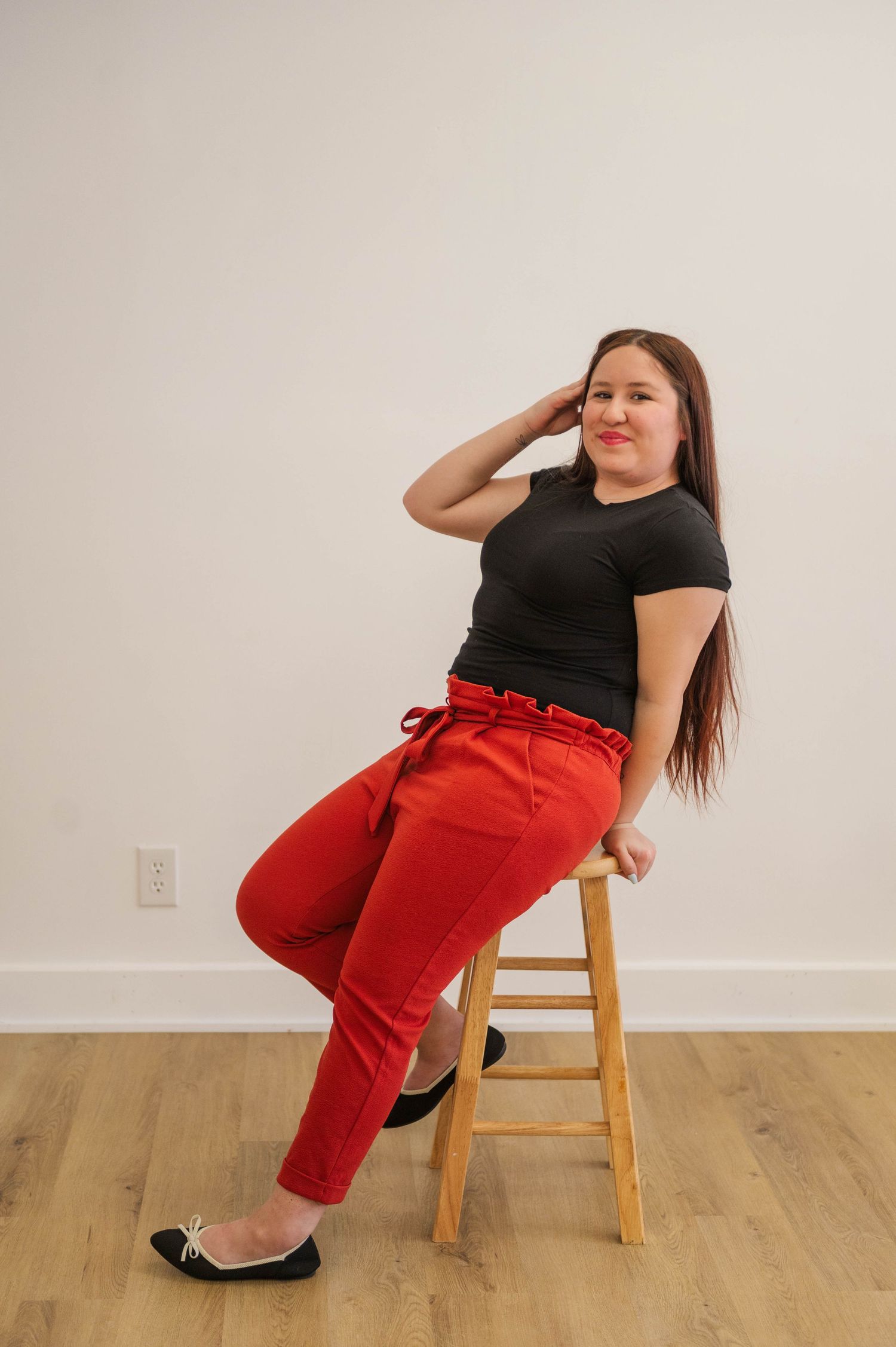 Brunette woman sits on wooden stool in black tee and red paperbag pants, smiling with hand near hair.