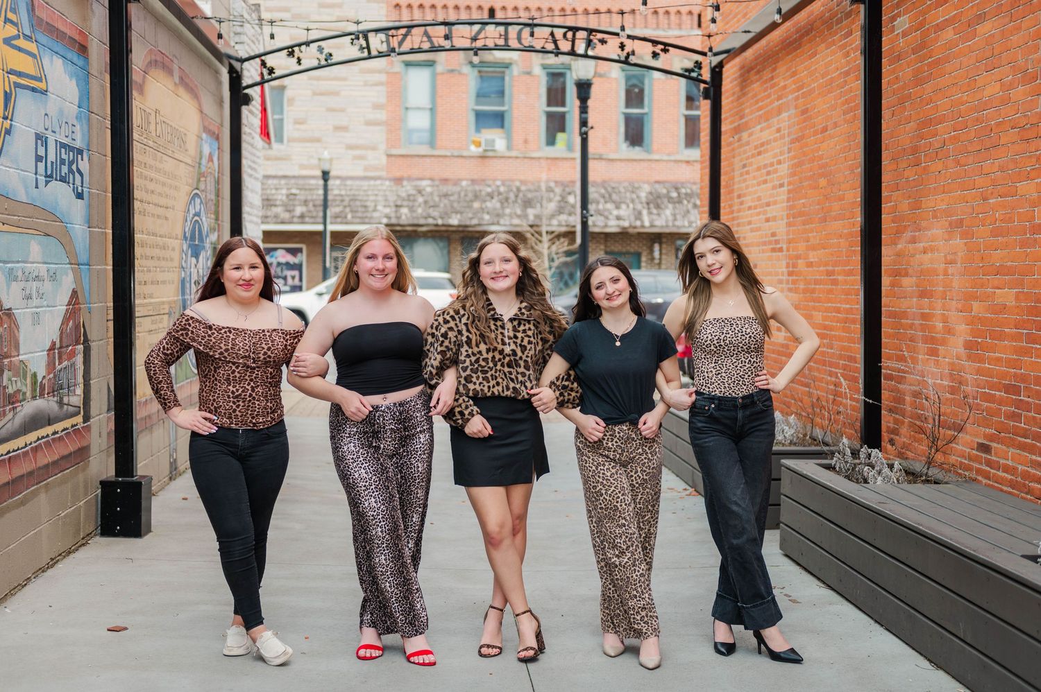 Five stylish women in animal print outfits pose together in a brick urban alleyway.