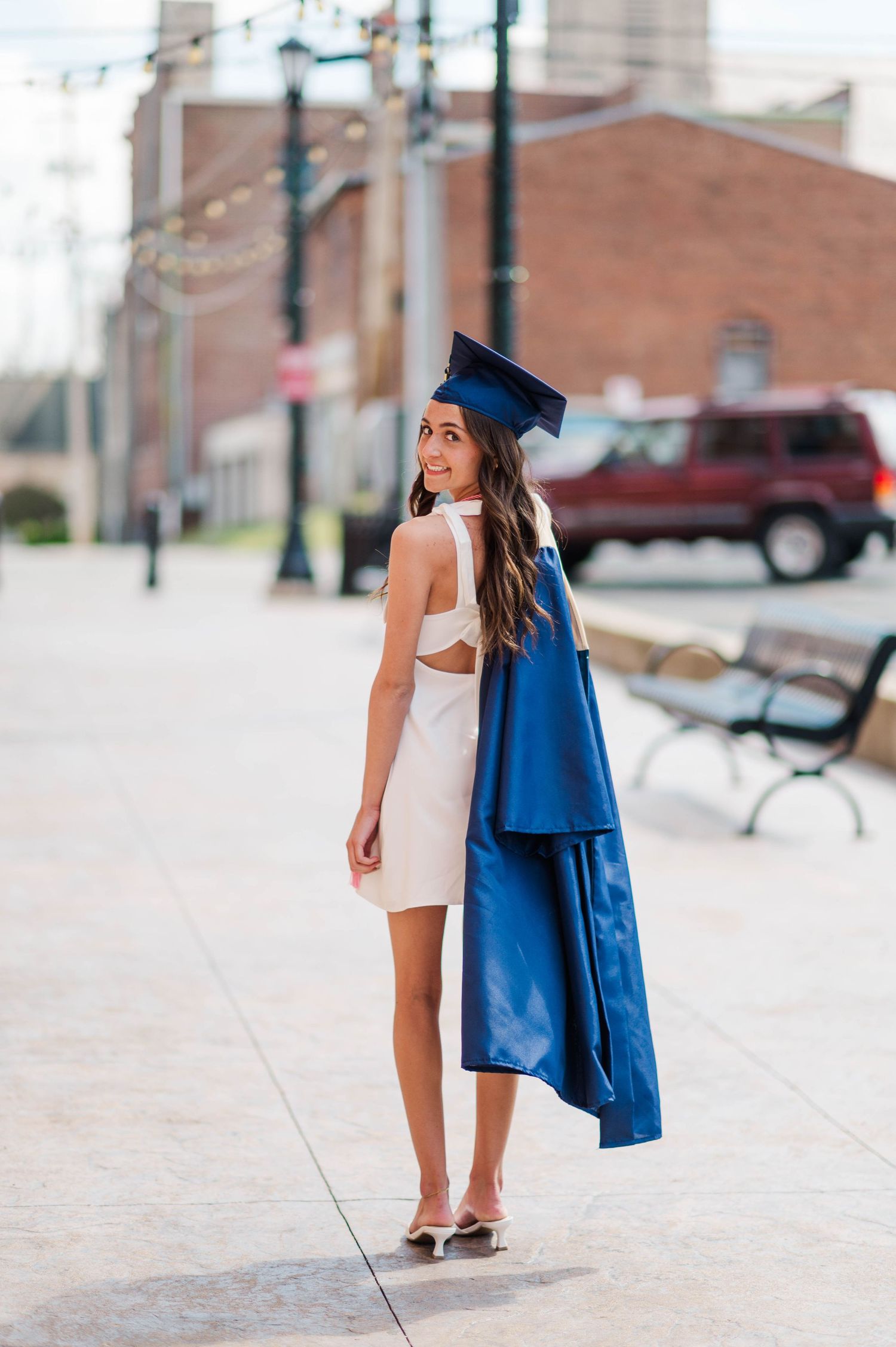 Brunette woman in white dress carrying navy graduation cap and gown on sunny urban walkway.