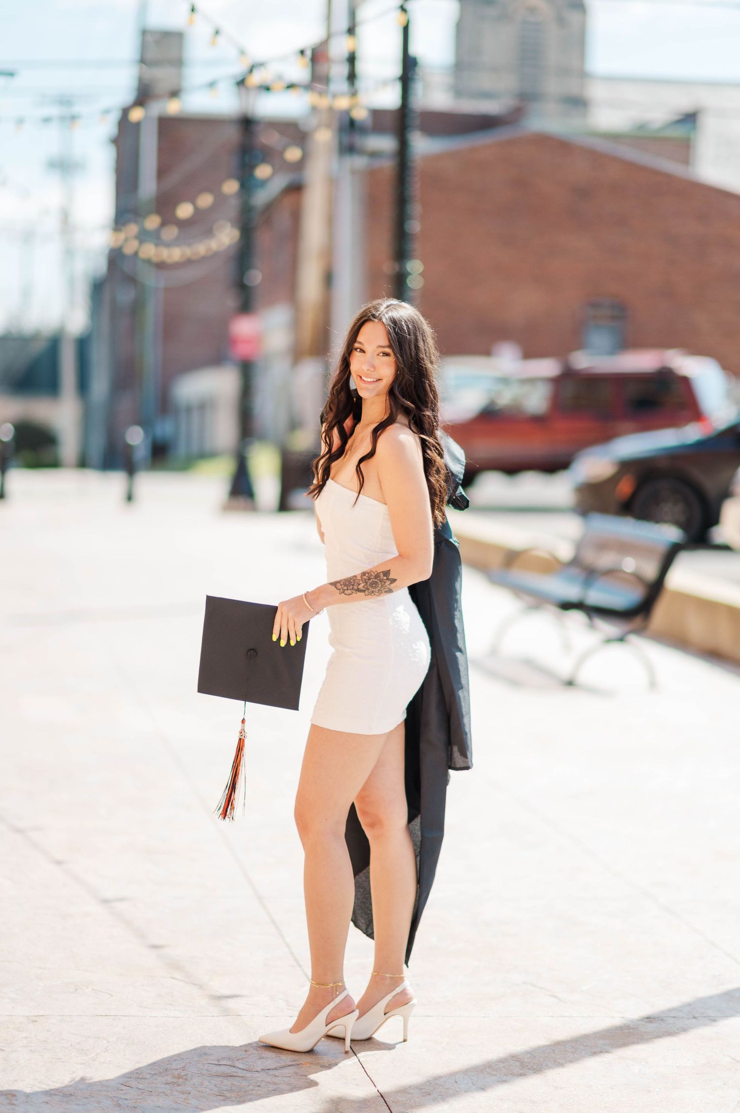 Stylish graduate in white dress and black gown holding cap with tassel on sunny urban street.