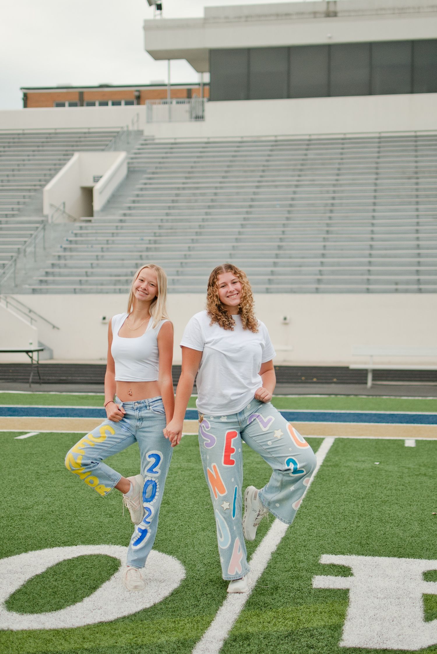 Two young women in white tops and painted graduation jeans pose on a football field with stadium bleachers in the background.